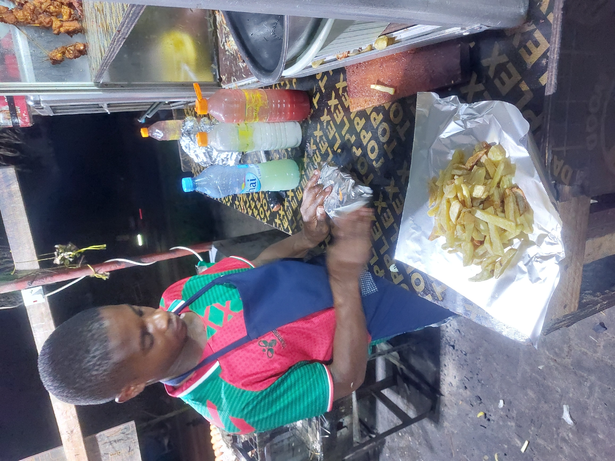 The image captures a street food vendor in Dar es Salaam, Tanzania. The vendor, a young man, is wrapping a portion of french fries in aluminum foil. He is positioned behind a wooden food stall. On the stall's surface, there are plastic bottles filled with colorful liquids. Also, the stall has a metallic tray. A pile of cooked skewers is visible at the stall's edge. The background indicates an urban setting with street lighting.