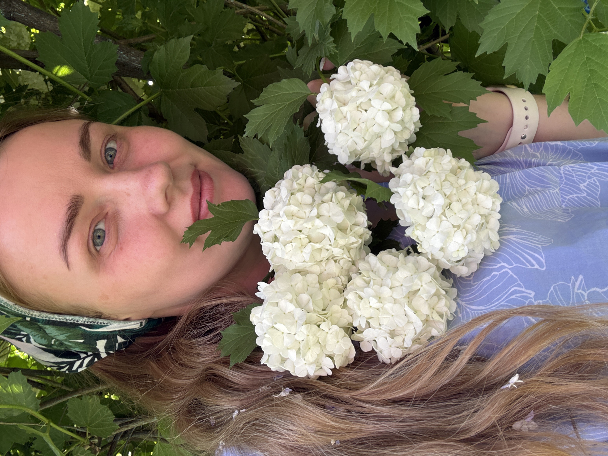 A fair-skinned woman with blonde hair is lying on her side amidst foliage and white flower clusters. She wears a green and white patterned headband and a blue patterned dress. Her right arm is visible, adorned with a white watch or bracelet. The background is filled with green leaves and branches. Small white flowers and petals are scattered on her hair. The image was taken in Chaikovsky, Russia.
