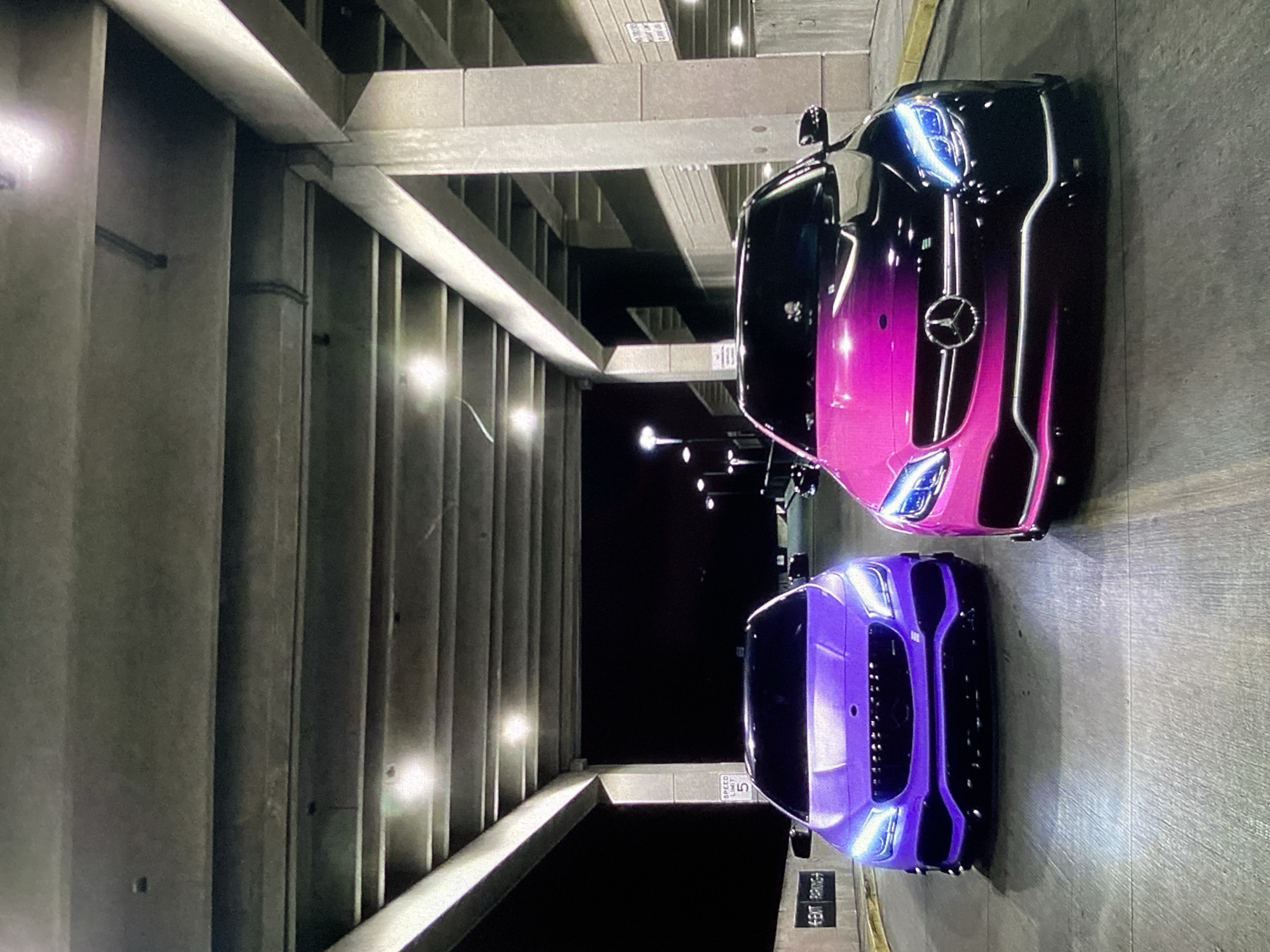 The image shows two luxury vehicles parked in an indoor parking garage. The vehicle closest to the viewer is a purple coupe with its headlights on, facing the camera. Behind it is a car that transitions from black to pink in color. The parking garage has exposed concrete beams, pillars, and ribbed walls. The lighting is dim, and there are overhead lights visible in the background. There are no people present in the image. There is no indication that the image was taken in Lhokseumawe, Indonesia. The location is simply an indoor parking garage.