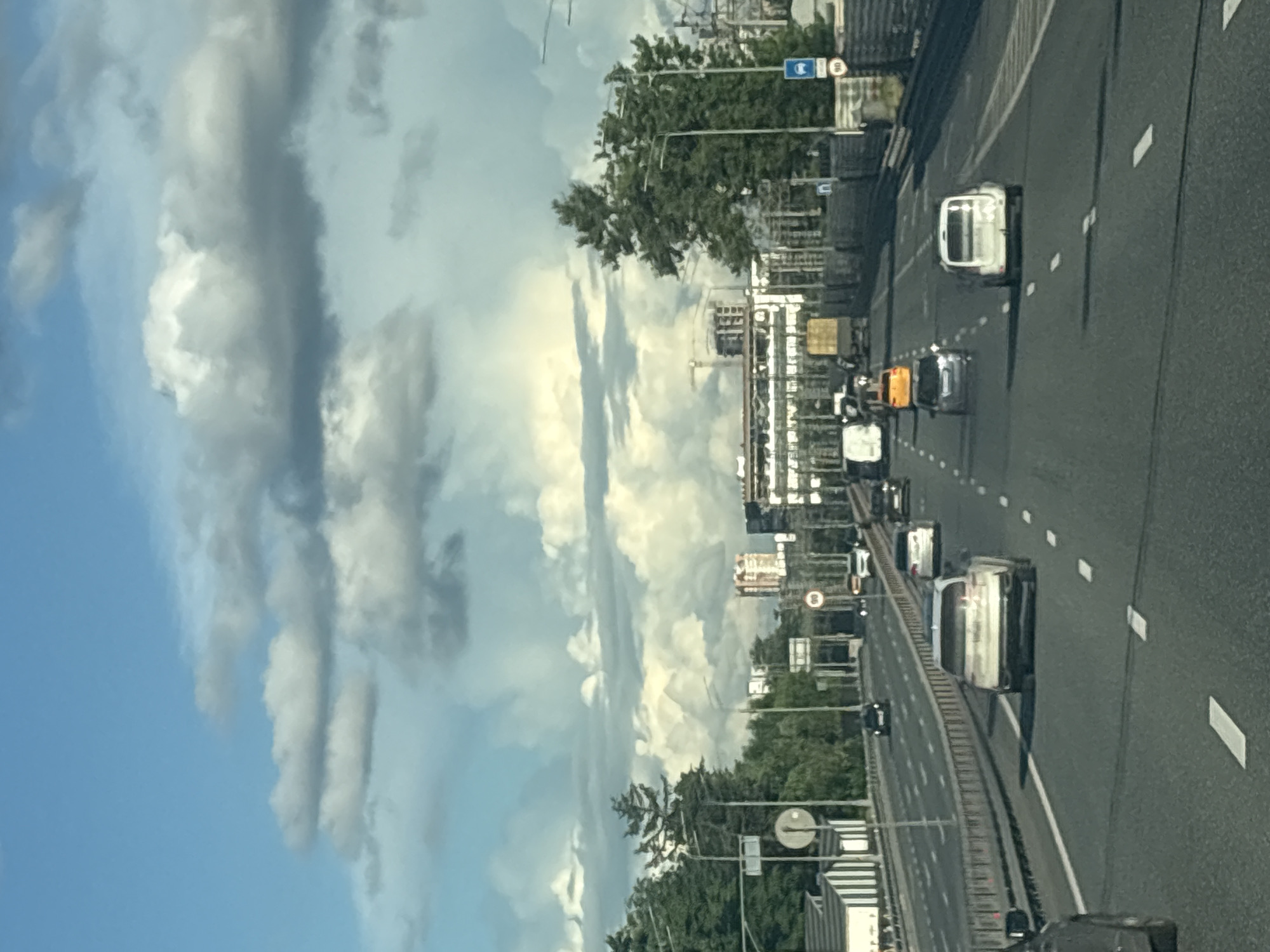 The image depicts a multi-lane highway with several vehicles driving in the same direction. The perspective is from a car, suggesting the viewer is a passenger or driver. The sky is partially cloudy with bright white clouds against a blue sky. On the left side of the highway, there are trees and buildings.
The vehicles on the road include a white van or camper, a yellow vehicle, a dark car, and several other cars further down the road. The road is paved and marked with white lines. There are also signs visible, including a circular sign with the number "80" inside, indicating a speed limit.
The overall scene suggests a daytime drive on a highway, possibly in an urban or suburban area given the presence of buildings and trees. The weather appears to be fair with some cloud cover. The image is taken from a slightly elevated angle, looking down onto the highway. No people are clearly visible within the vehicles. The image quality is somewhat grainy, and the orientation is sideways.