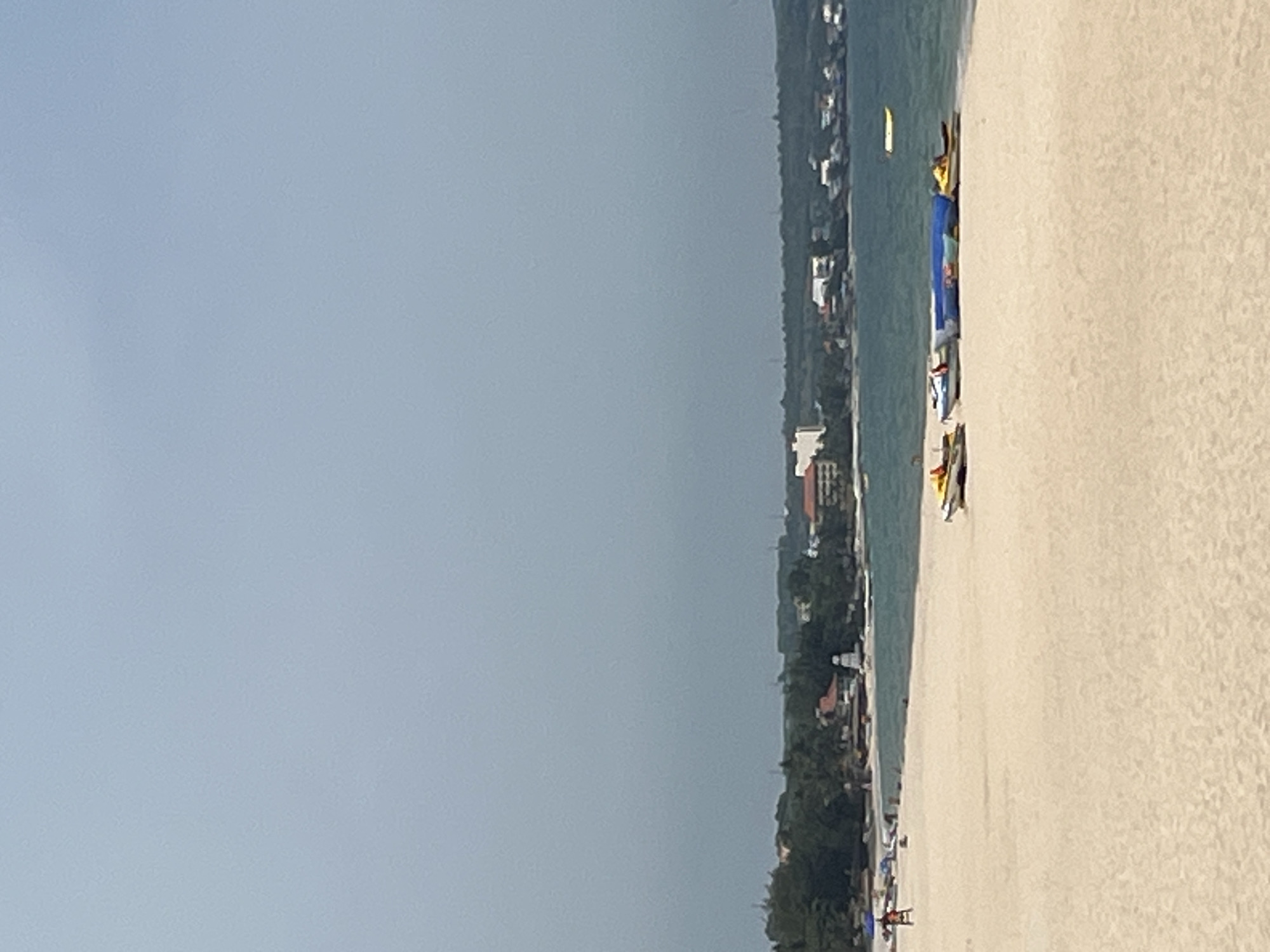 A beach scene is depicted with a calm sea under a hazy sky. In the foreground, a sandy beach stretches across the frame. Several watercraft, including jet skis and what appear to be small boats, are parked on the sand near the waterline. Some of these are covered with blue tarps. In the distance, a line of trees and buildings marks the coastline, indicating a populated area. A few umbrellas and people are visible on the beach, suggesting a tourist or recreational setting. A yellow buoy floats in the water to the right. The overall impression is a peaceful beach day in a coastal Vietnamese town, likely Phu Thuy Ward.
