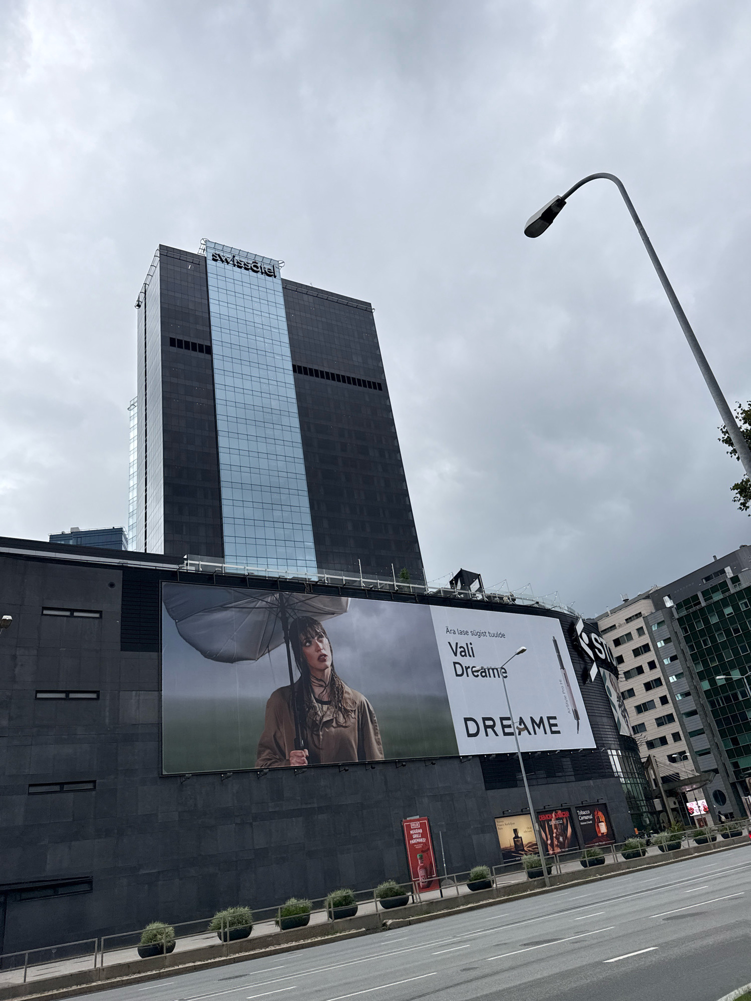 The image shows a street view in the city of Tallinn, Estonia, on a cloudy day. In the foreground, a street and sidewalk are visible, with several potted plants lining the sidewalk. Behind the sidewalk, a large, dark gray building dominates the scene. Attached to the side of this building is a massive billboard featuring a woman holding an umbrella. The billboard text includes "Ara lase sügist tuulde Vali Dreame" and "DREAMЕ". Above this building, a tall, modern skyscraper with dark and glass panels rises into the sky. The skyscraper has a prominent "SWISSOTEL" sign at its peak. To the right of the skyscraper, another modern building with a grid-like facade is visible. A streetlamp with a curved arm is positioned in the upper right quadrant of the image. The sky is overcast and gray.