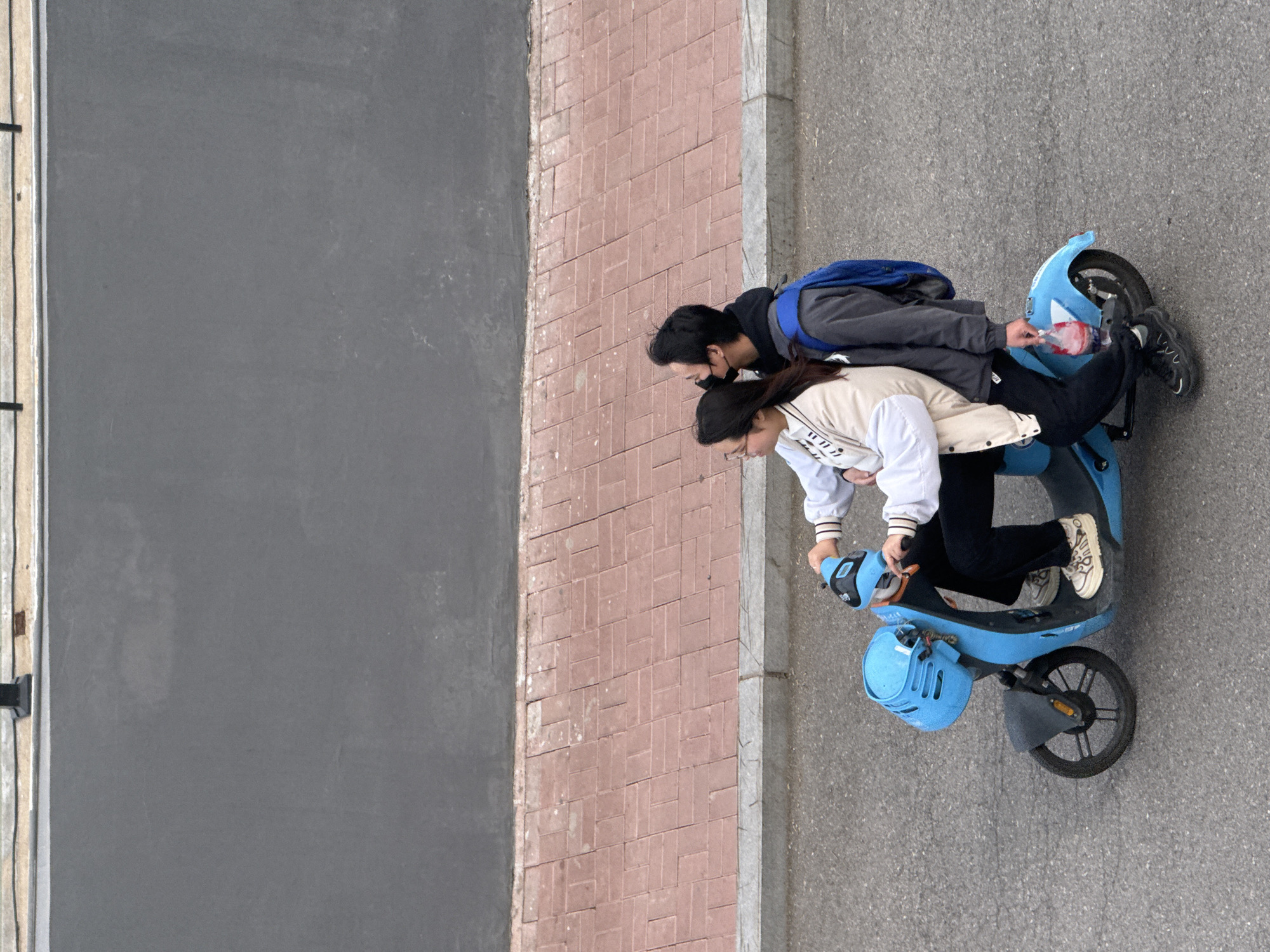 Aerial view of two people on a light blue electric scooter on a gray asphalt surface in Zhongtun, China. A person in a white jacket and dark pants operates the scooter, and a second person with a blue backpack rides as a passenger. The scooter has a basket on the front and a helmet hanging on the basket. A gray concrete sidewalk is separated from a brick surface by a gray curb. To the left of the brick surface is a gray wall. The passenger is carrying a plastic bottle with a red and white label.
