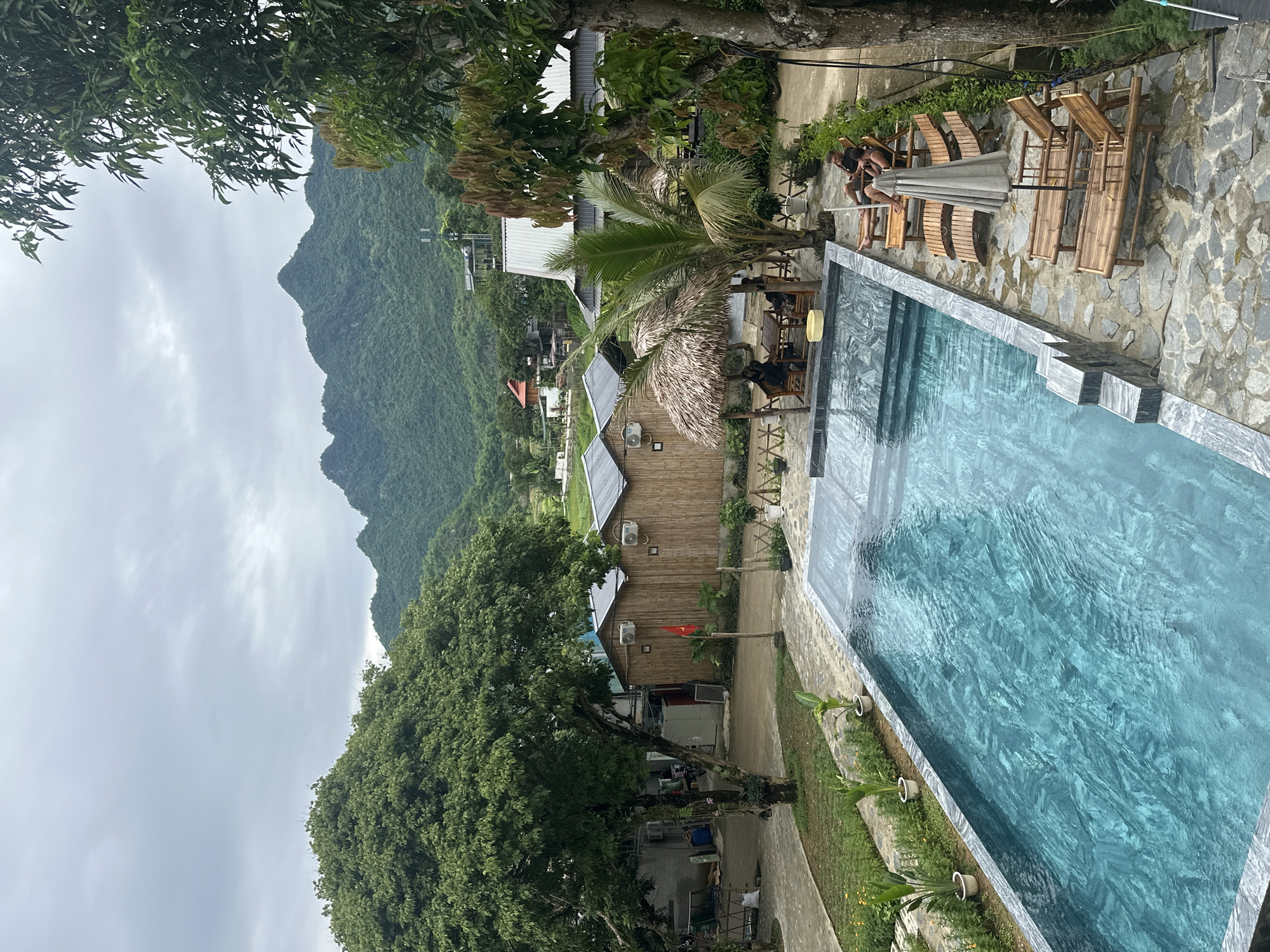 This image captures a serene resort scene in Mai Chau, Vietnam, with a prominent swimming pool in the foreground. The pool, filled with clear turquoise water, features a stepped entry and a stone tile border. Surrounding the pool, a stone patio is furnished with several empty wooden lounge chairs and one umbrella, though no one is currently using them. Further back, a thatched-roof gazebo shelters a table where two individuals are seated. Beyond the pool area, traditional bamboo huts and modern structures with metal roofs are visible, hinting at the resort's accommodation. In the distance, lush green mountains dominate the horizon under a cloudy sky, framing the entire landscape. A large tree stands to the left, casting shadows and adding to the natural beauty of the setting. The overall atmosphere suggests a tranquil retreat in a rural mountainous region.