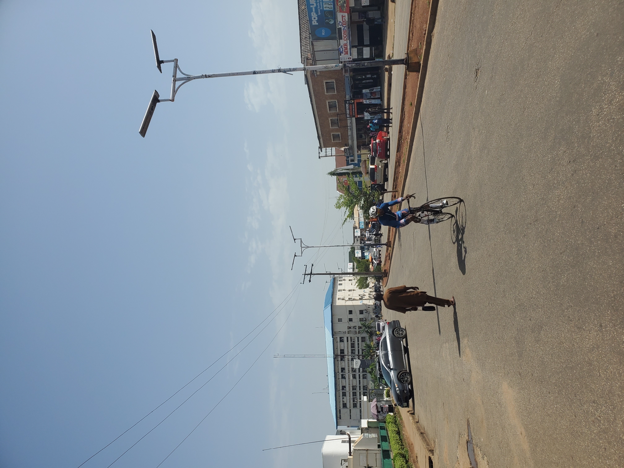 The image depicts a street scene in Jos, Nigeria. In the foreground, a person is riding a bicycle on a paved road. To the left of the cyclist, another person in traditional brown attire is walking away from the camera. Several cars are parked and driving on the road. In the background, there are buildings lining the street, including a large white building with a blue roof. Overhead, power lines are visible against a clear blue sky. The environment appears to be urban, with streetlights and shops along the road.