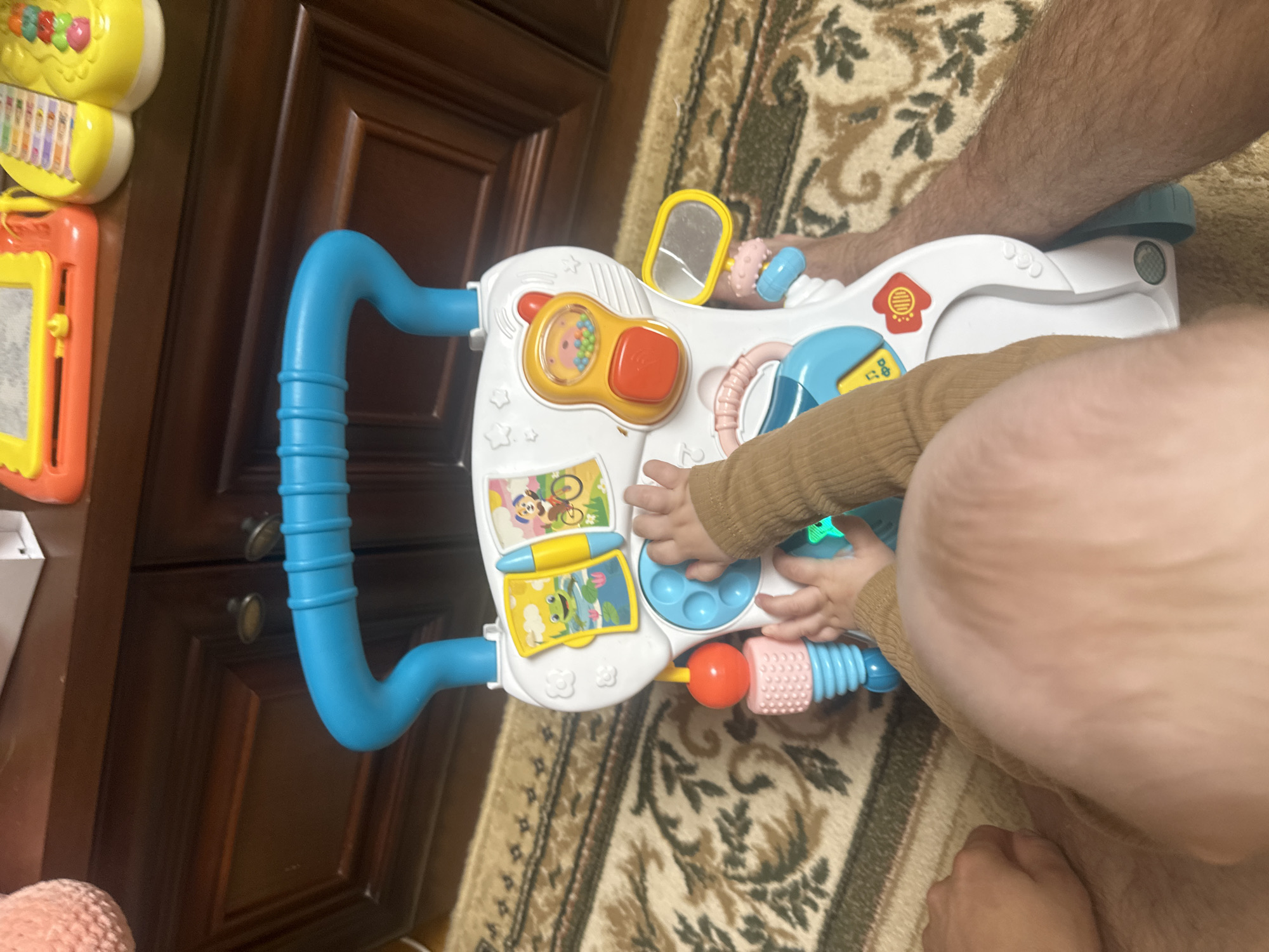 A baby is interacting with a white and blue baby walker. The baby is wearing a brown long-sleeved shirt and its hands are on the walker's interactive panels. The walker has various colorful buttons, a mirror, and decorative elements. The scene is set indoors on a patterned rug, with a wooden cabinet visible in the background. The setting is likely a home in a city, as there is no information to suggest it is in Загородный, Russia.