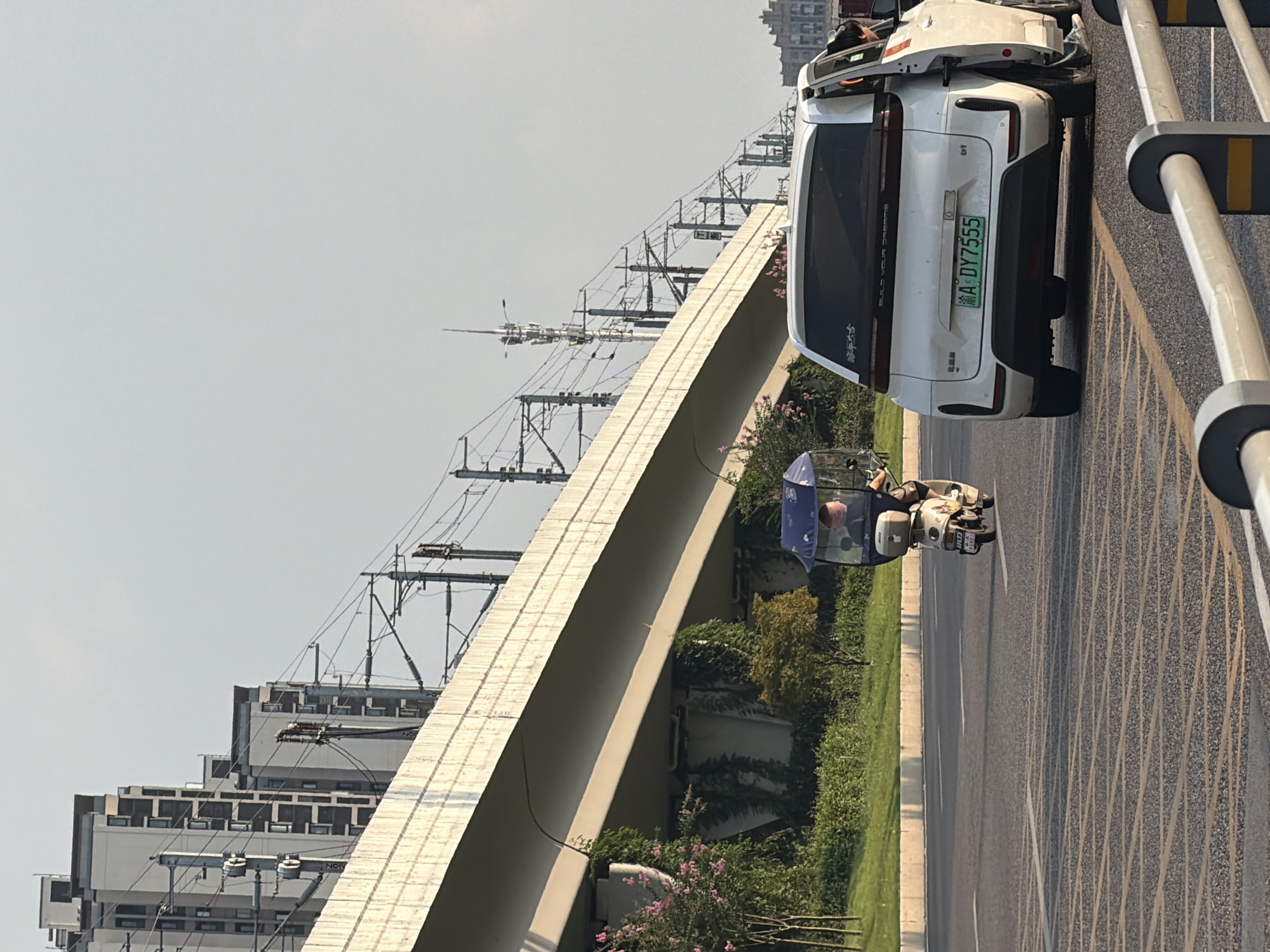 In the city of Jiangjin, China, a white electric car with its passenger door ajar is positioned on the right lane of a multi-lane asphalt road. A license plate reading "A DY7555" is visible on the rear of the car. Ahead of the car in the left lane, a person rides a three-wheeled scooter covered with a blue canopy, which has a dark brown box mounted on the rear. The scooter also displays a license plate. The road is lined with a metallic guardrail in the foreground. In the background, a raised concrete structure, likely an elevated railway or highway, spans across the scene. Overhead, power lines and utility poles are strung along the structure. The environment features a clear sky and a few trees and bushes bordering the road. In the far background, several tall buildings with repeating window patterns are visible, indicating an urban setting.