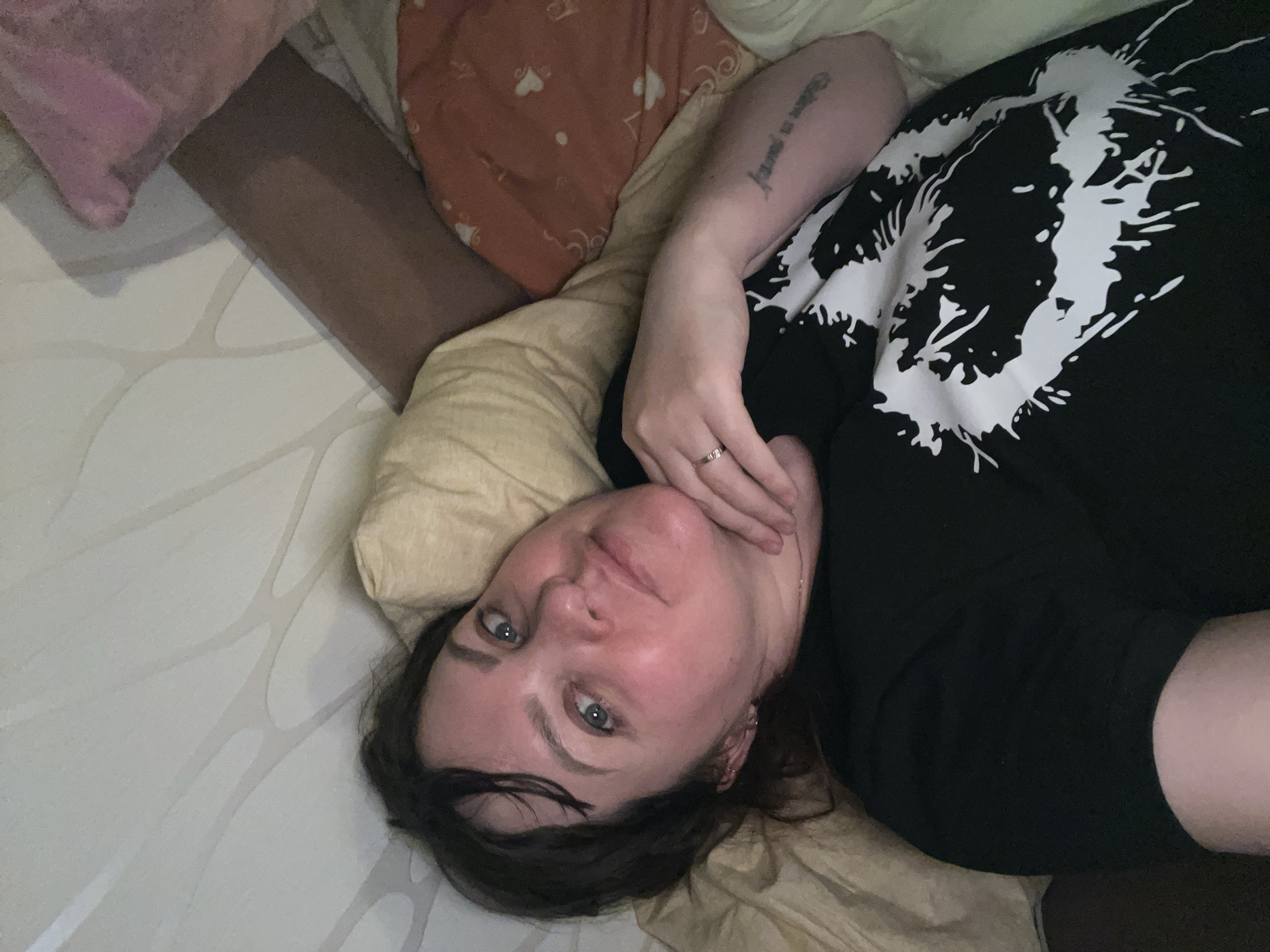 A woman is captured in a close-up, high-angle shot, lying on her back on a bed in what appears to be an indoor setting in Perm, Russia. She is looking directly at the camera with a calm expression, her eyes a striking blue-green. Her dark hair is casually spread around her head on a light-colored pillow.
She is wearing a black t-shirt featuring a prominent white, abstract, splattered design on the chest. Her right hand is resting near her chin, revealing a simple ring on her ring finger. A tattoo, consisting of text in a script font, is visible on her inner left forearm; the words "Believe in yourself" can be deciphered.
The bed is covered with a light-toned sheet that has a subtle, organic-line pattern. Several pillows are arranged around her, including a pink one, an orange one with a faint heart pattern, and a plain beige pillow under her head. The lighting suggests an indoor environment, possibly a bedroom, with no strong indications of the time of day or specific weather. The overall impression is one of relaxed repose or a casual self-portrait.