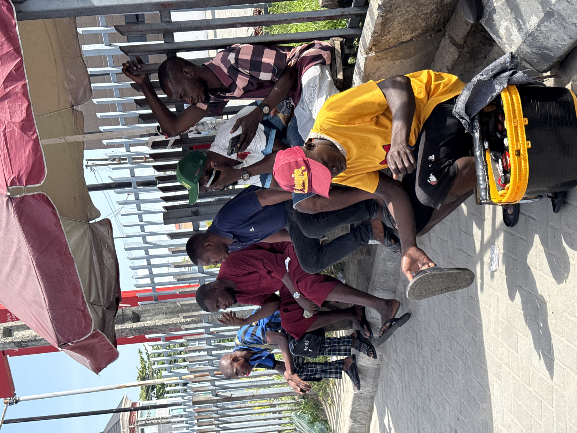 A group of six young men are seated closely together against a metal railing and a concrete wall, along a paved walkway in Ado, Nigeria. They appear to be engaged in watching something on their mobile phones, as each is looking down at a device in their hands. The men are dressed in a variety of casual clothing including t-shirts, shorts, trousers, and sandals. One wears a pink baseball cap, another a green cap, and another a yellow t-shirt. An open toolbox sits near one of the men, its contents visible. A red and beige umbrella provides partial shade.