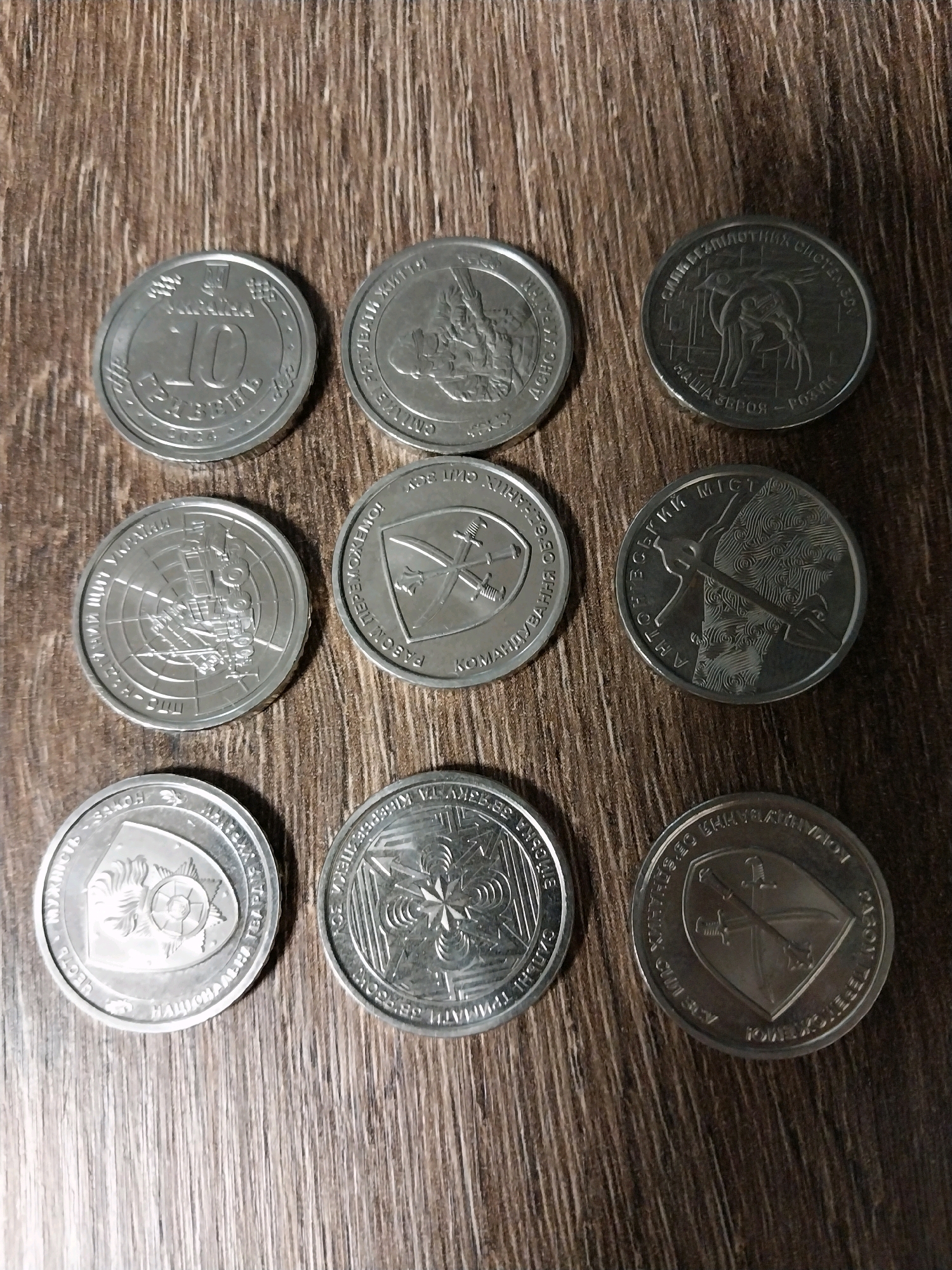 Nine coins are laid out on a wooden surface. The coins appear to be of Ukrainian origin, based on the Cyrillic inscriptions and iconography. The wooden surface has a prominent grain pattern. From top left to bottom right, the coins are:
1. A coin with the number "10" prominently displayed, along with other inscriptions.
2. A coin featuring a mythological or allegorical scene with figures.
3. A coin with an image of a bird-like creature and inscriptions that appear to read "СИЛА БЕЗПІЛОТНИХ СИСТЕМ".
4. A coin with a design featuring a ship or vessel within a circular pattern.
5. A coin depicting crossed swords within a shield, with inscriptions.
6. A coin with an image of what appears to be a crossbow and stylized hair or clouds.
7. A coin with a shield-like emblem and inscriptions that include "ЗАКОН".
8. A coin with a star-like pattern within a circular border.
9. A coin featuring a shield with crossed swords and an inscription.