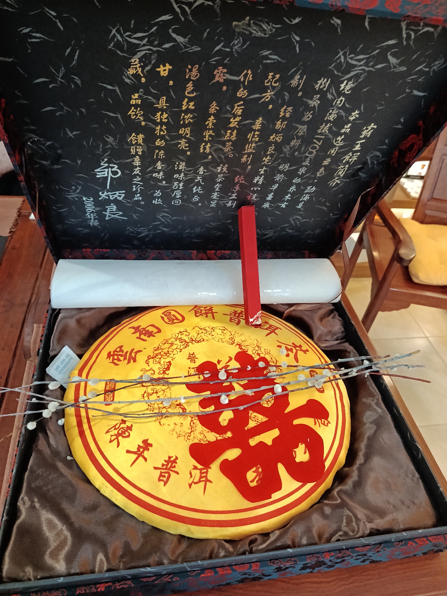 The image shows a box containing a round, yellow tea cake with red Chinese characters on it. A red item and a white cylindrical object are next to the tea cake. Pussy willow branches are placed on top of the cake. The inside lid of the box is black with gold-colored Chinese calligraphy written on it. The tea cake and other objects are set on brown fabric. A chair with a yellow cushion is in the background. There is no indication of the location being Moscow, Russia.