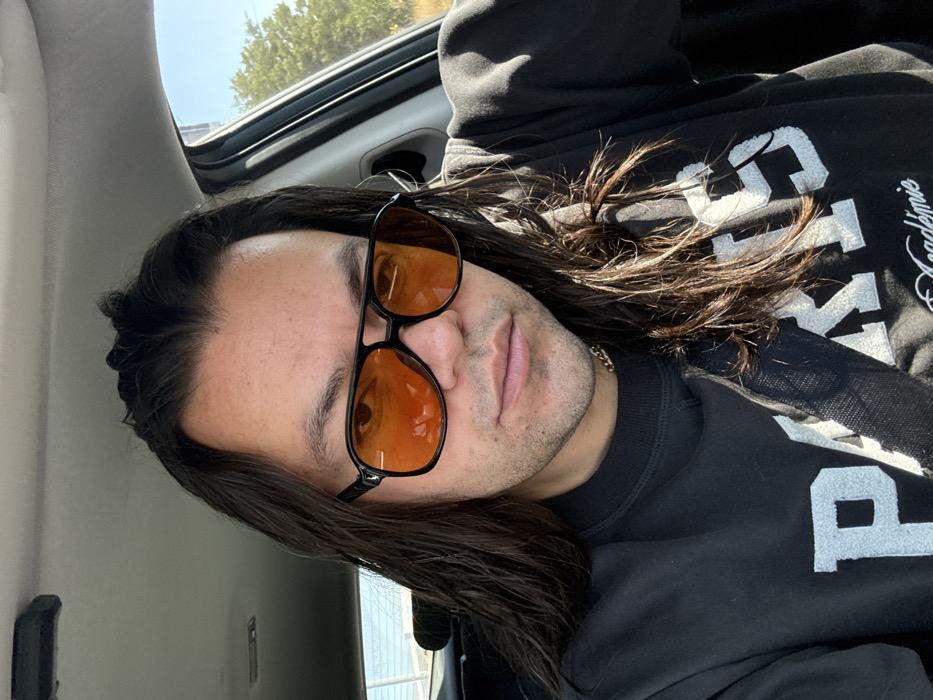 An adult individual, identified as male, with long, dark brown hair and light facial stubble, is depicted seated in the driver's seat of a vehicle. He wears black-framed aviator-style sunglasses with orange-tinted lenses and a thin gold-colored chain around his neck. His attire consists of a black long-sleeved crew-neck sweatshirt with "PARIS" in white textured lettering and "Académie" written below it. A black seatbelt is fastened across his chest, tautly secured. The individual is looking directly towards the camera. Sunlight enters from the right, illuminating the subject and interior. Through the vehicle's windows, a clear blue sky and green foliage are visible in the background, consistent with an outdoor, daytime environment. The specific location in Barrio de Nuevo León, Mexico, cannot be determined from the visual evidence within the image.