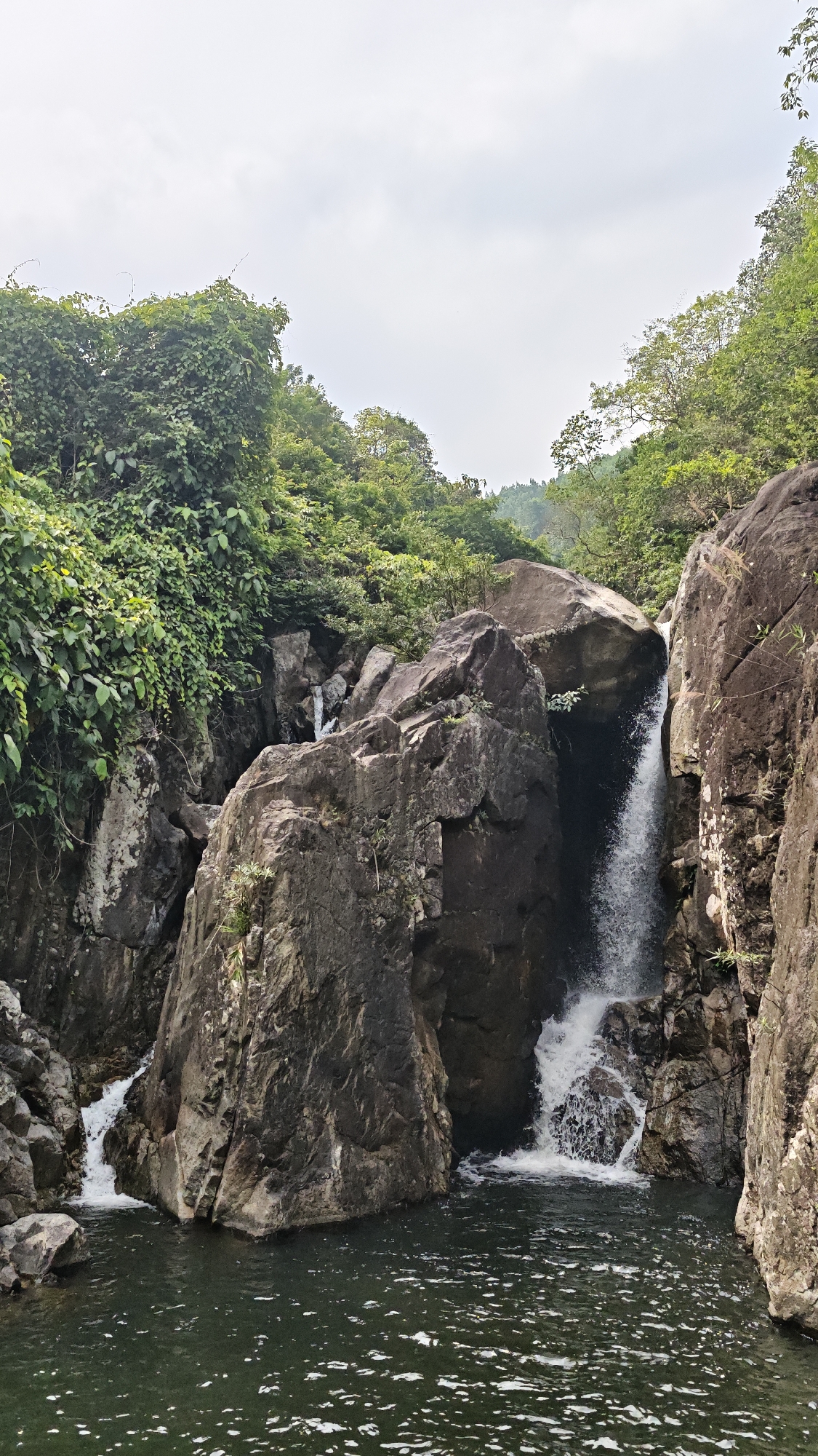 The image shows a waterfall flowing into a pool surrounded by large rocks and lush greenery. Trees and vegetation cover the rocks and the area surrounding the waterfall. The sky is overcast. The location is in the city of Đà Nẵng, Vietnam.