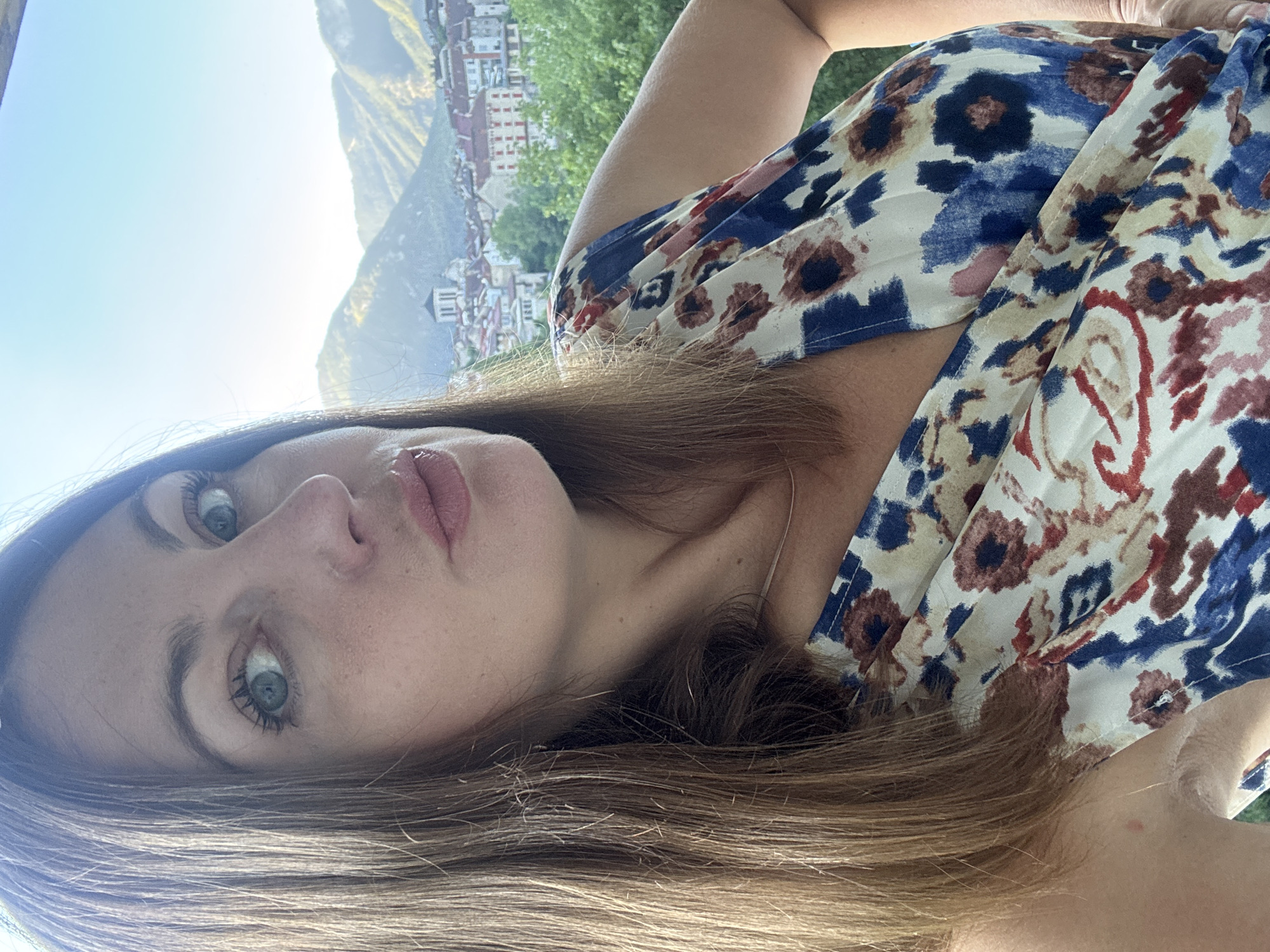 The image is a selfie of a woman with long brown hair and blue eyes, wearing a floral print dress. She is outdoors, with a view of a town nestled in a valley with mountains in the background. The town appears to be Saint-Claude, France, characterized by its red-tiled roofs and a prominent church tower. The woman's pose suggests she is leaning forward, looking at the camera. The lighting indicates it is daytime, possibly late afternoon, given the angle of the sun casting light on the mountains. The overall impression is a casual snapshot taken with a scenic backdrop.