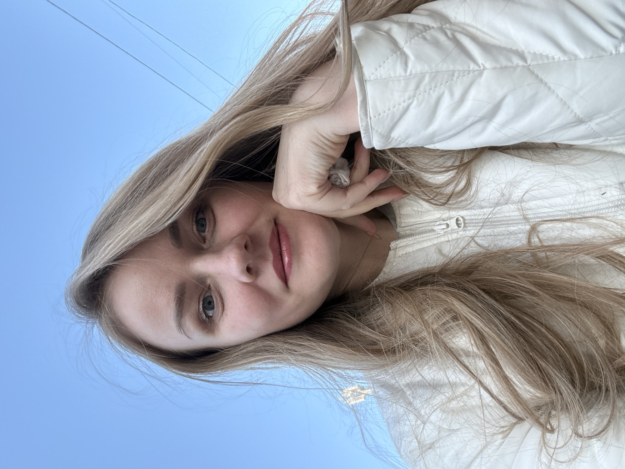 A woman with long blonde hair, wearing a white quilted jacket, poses for a selfie against a backdrop of a clear blue sky. Power lines are visible in the top portion of the frame. The location is Chaikovsky, Russia.