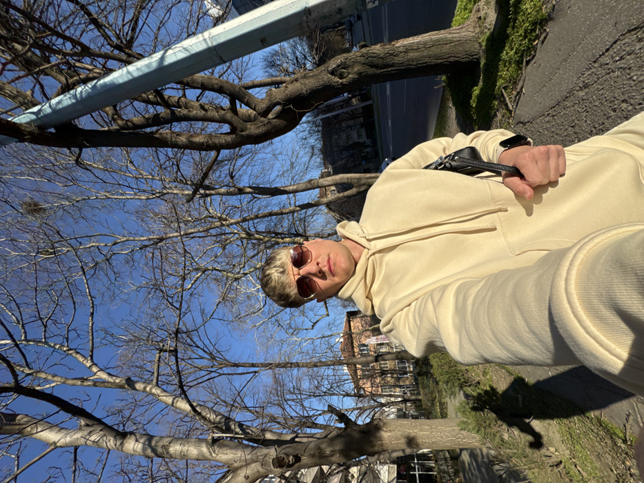 A male individual is depicted in a selfie taken outdoors on a sunny day in Plovdiv, Bulgaria. The subject, positioned centrally, has light-colored hair and wears aviator-style sunglasses with amber-tinted lenses. He is dressed in a cream-colored hooded sweatshirt. His extended left arm holds the camera, while his right hand holds a small, black shoulder bag. A dark wristwatch is visible on his left wrist. The background features a clear blue sky and several bare-branched deciduous trees, indicative of late winter or early spring; a bird's nest is visible in an upper branch of one tree. A paved sidewalk and sparse ground vegetation are in the foreground, with the subject's shadow prominent on the left. In the mid-ground, urban buildings are visible, including one with a reddish-brown tiled roof and light-colored facade. A light-colored utility pole and a street with parked vehicles are visible on the right.