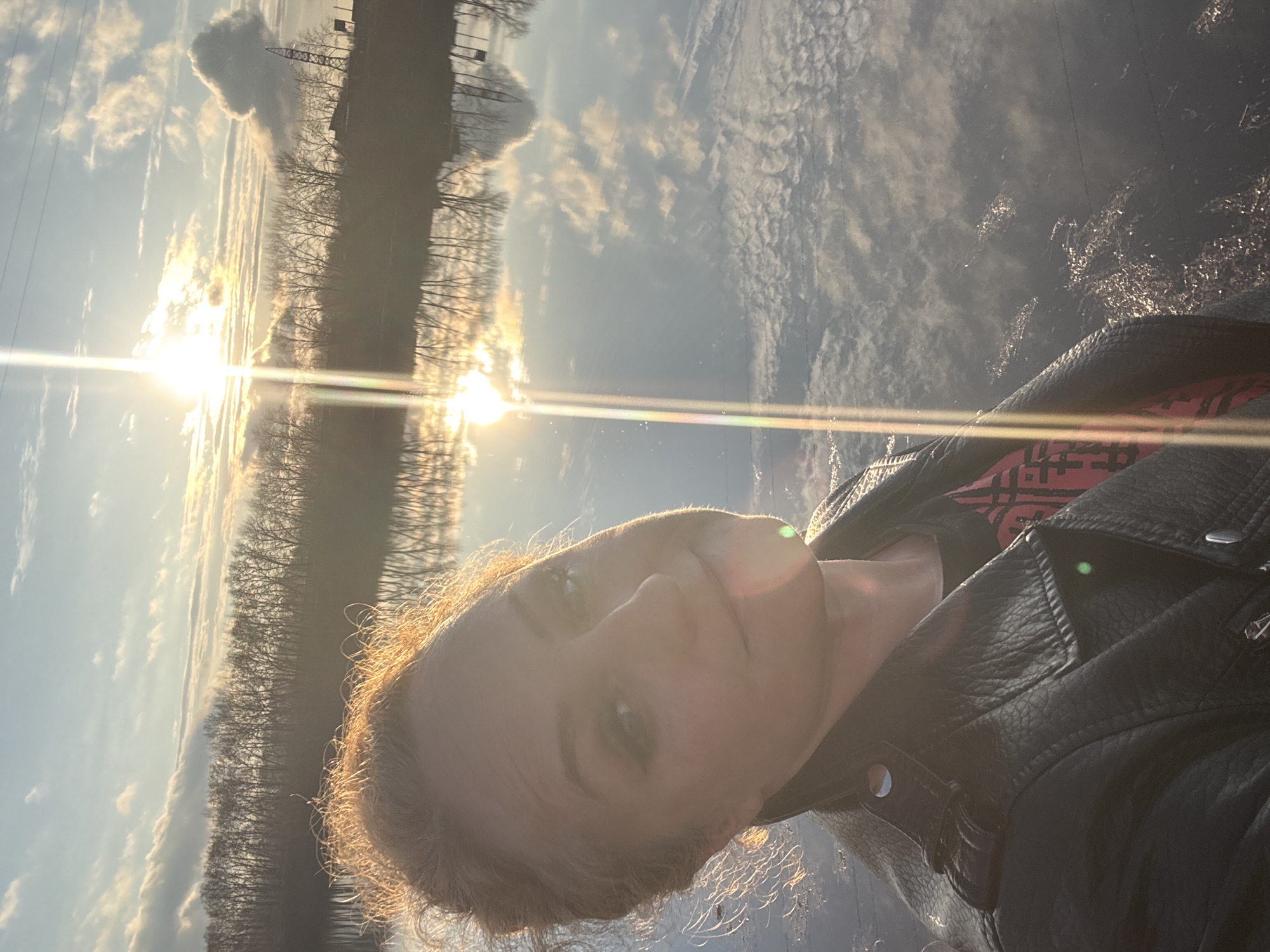 A selfie captures a woman outdoors, facing the camera with a calm expression, possibly a faint smile. Her light-colored hair is back-lit by a bright, low sun, creating a luminous halo effect around her head. She wears a black leather jacket over a red garment with indistinct dark patterns. A prominent lens flare from the sun diagonally crosses the image.
The setting, likely near Chaikovsky, Russia, features a wide body of water in the background, reflecting the bright sky and scattered clouds. A line of bare, deciduous trees stands on the distant shore, their silhouettes visible against the glowing sky and their reflections mirrored in the water. The water's surface shows subtle ripples and sparkling highlights from the sunlight. The sky above has scattered clouds, illuminated by the warm light of what appears to be either sunrise or sunset. The natural light suggests a clear day. The scene evokes a serene and reflective atmosphere during the golden hour. No decipherable text is visible.