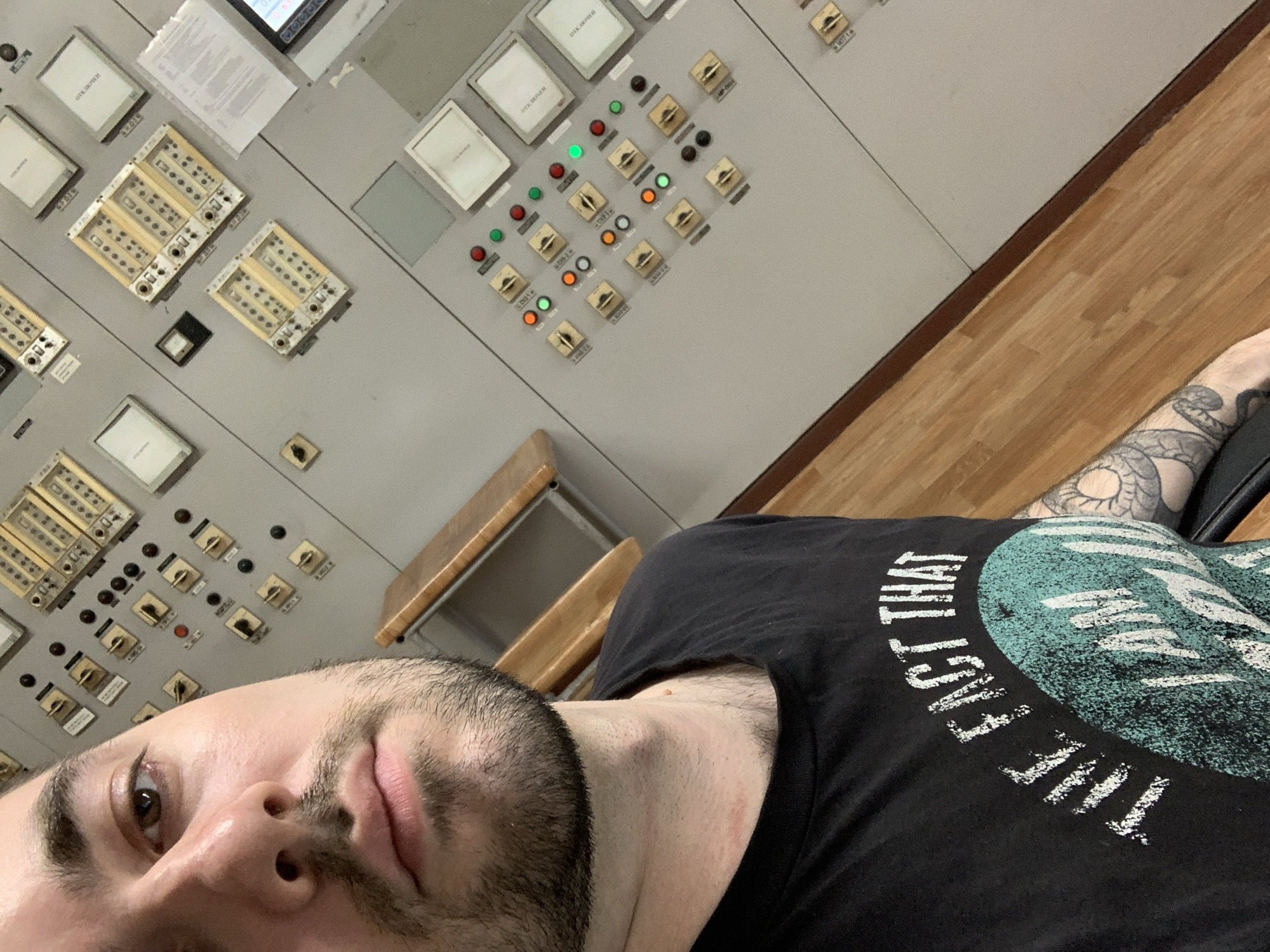 A man is taking a selfie in what appears to be an industrial control room in Chaikovsky, Russia. The image is taken at an oblique angle, with the man's face and upper body dominating the foreground, while a large control panel and a wooden floor occupy the background.
The man has a beard and dark hair, with a neutral expression as he looks towards the camera. He is wearing a black t-shirt with white distressed text that partially reads "THE FACT THAT I AM". On his left arm, visible on the right side of the image, is a detailed tattoo of a snake or serpentine creature.
The background features a grey control panel or console, densely packed with various operational components. These include multiple square displays, some labeled "OTK/OKHOEM" (likely Russian text), and others with less legible alphanumeric labels. There are numerous toggle switches or buttons, accompanied by indicator lights, with several green, red, and orange lights illuminated. Various rectangular modules with arrays of small sockets or indicators are also part of the panel. A piece of paper with printed text is affixed to the upper section of the control panel. The floor is made of light-colored wooden planks, suggesting an interior industrial or operational setting. No specific activity other than taking the selfie is observed.