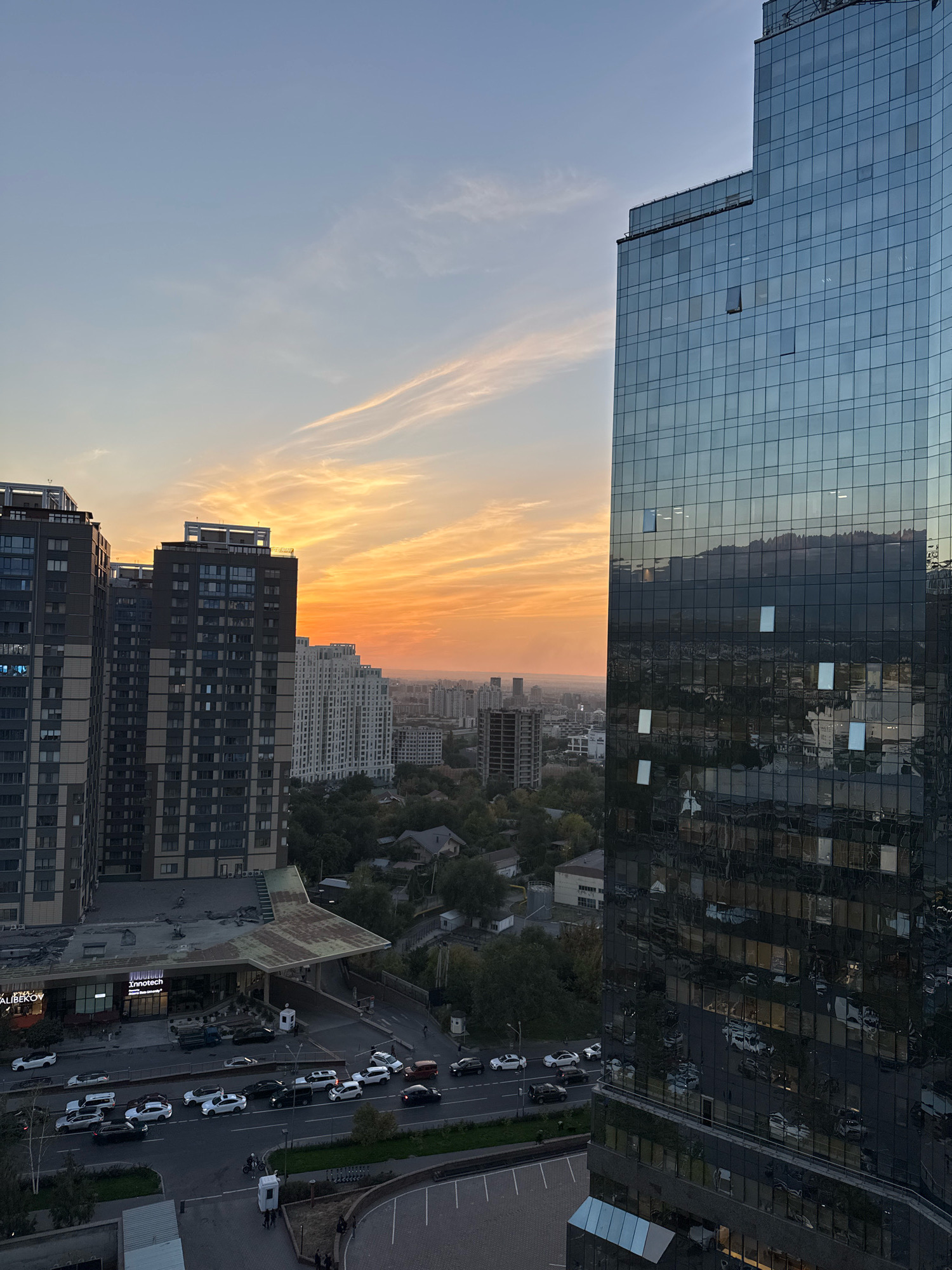 The image depicts a city scene in Almaty, Kazakhstan, likely at dusk, with a vibrant sunset coloring the sky. In the foreground, a modern, glass-facaded skyscraper dominates the right side of the frame, reflecting the sky and surrounding buildings. To the left, a cluster of residential buildings with a mix of dark and light facades are visible, some featuring balconies and numerous windows. Below these buildings, a street is lined with parked and moving vehicles, including sedans, SUVs, and trucks. A low-rise commercial building with a distinctive curved roof is situated between the residential buildings and the street. In the background, the cityscape extends towards the horizon, with more buildings and trees visible, and a faint mountainous silhouette can be discerned in the distance under the colorful sky. The overall impression is one of an urban environment transitioning into evening, with a focus on modern architecture and city infrastructure.