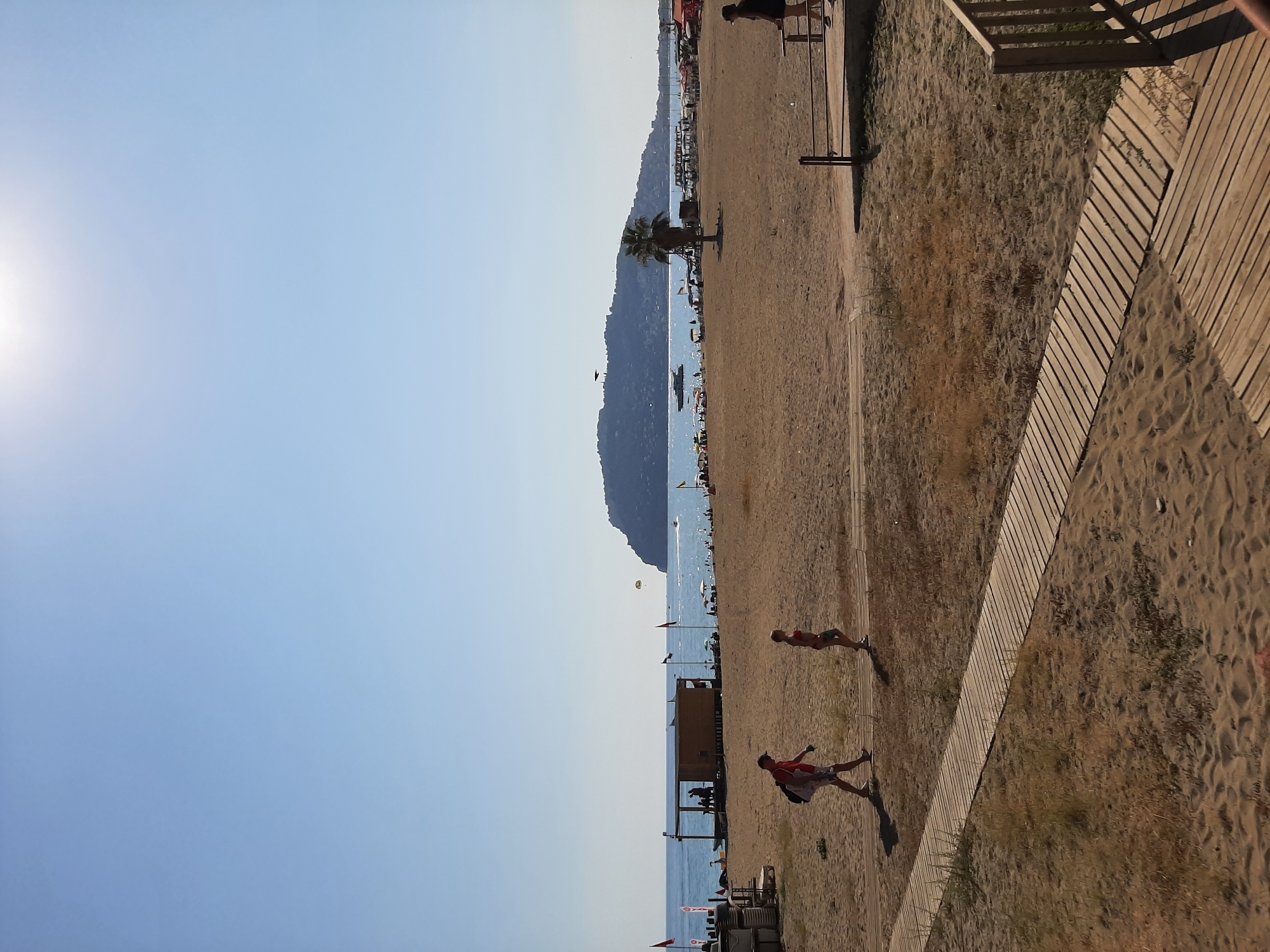 The image depicts a bright, sunny day at a beach in Alanya, Turkey, likely Cleopatra Beach given the prominent hill in the background. The foreground features a sandy beach with a wooden boardwalk. Two individuals are walking on the boardwalk; one is a man in a red shirt and shorts carrying a bag, and the other is a younger person in a red bikini. In the midground, there is a small wooden structure on the beach, possibly a beach bar or changing room, with people visible inside and nearby. Red flags are planted in the sand. The sea is calm and blue, with several boats visible on the water. Further in the distance, a large, green, hilly peninsula rises from the sea, characteristic of the Alanya landscape. The sky is clear and bright blue with the sun shining from the upper right corner, creating a lens flare effect. The overall scene conveys a relaxed beach atmosphere with a few people enjoying the sunny weather.