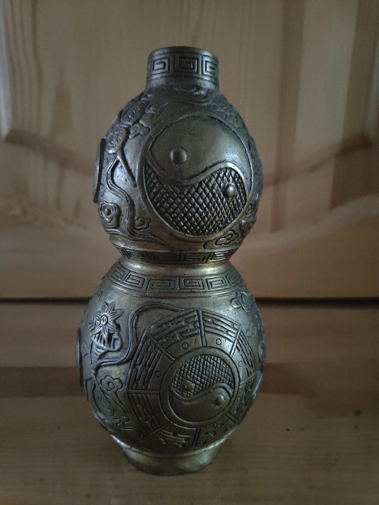 A close-up, eye-level shot shows a decorative, metallic gourd-shaped object with intricate carvings. The object is positioned centrally and appears to be made of brass or a similar metal, with a weathered, antique finish. The top portion of the gourd features a narrow neck and a rounded body decorated with a yin and yang symbol, surrounded by a textured grid pattern and stylized clouds. The bottom portion of the gourd is wider and also adorned with a yin and yang symbol, along with what appears to be a floral motif and more cloud-like designs. The object rests on a flat, wooden surface, likely a table or shelf. The background consists of vertical wooden panels, providing a warm, neutral backdrop. The lighting is soft and natural, casting subtle shadows and highlighting the textured details of the object. The overall scene is still and focused on the artifact. The location is Unknown, Unknown.