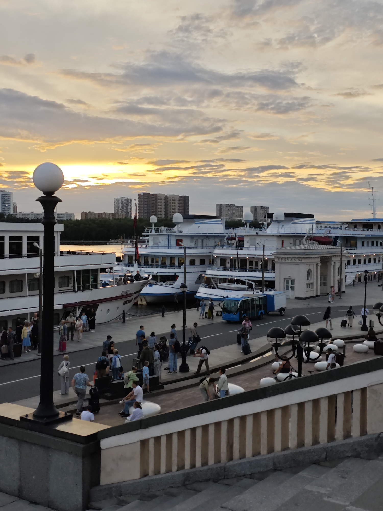 In Khimki, Russia, a sunset casts warm hues across a cloudy sky over a waterfront promenade. The scene depicts a marina with several large white passenger ships docked. People are visible on the promenade, some walking with luggage, suggesting travel or arrival/departure. A small blue shuttle bus is parked near the ships. A decorative lamppost with a spherical light fixture stands prominently in the foreground. Buildings and trees line the distant shore. The overall atmosphere suggests a bustling travel hub during the evening hours.