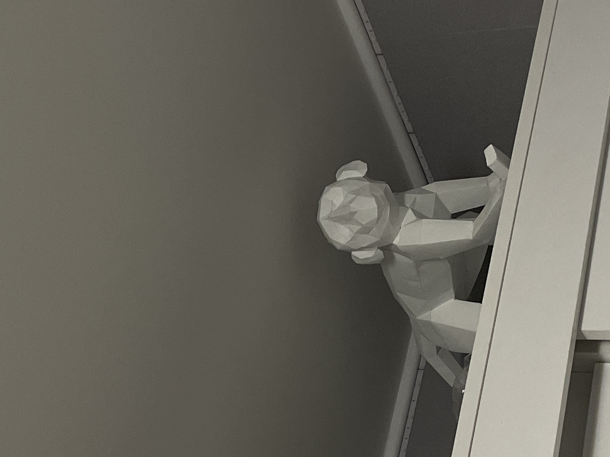 A low-angle, close-up shot shows a white, polygonal sculpture of a person in a climbing pose. The sculpture is made of geometric shapes, giving it a minimalist, abstract appearance. The figure is positioned on the right side of the frame, clinging to a white vertical element that appears to be a wall or pillar. The body of the sculpture is contorted, with one arm extended upwards and grasping the surface, and the legs bent as if in mid-climb. The head is turned away from the viewer, with facial features simplified into geometric planes.
The background is a muted gray, creating a stark contrast with the white sculpture and the white vertical element. The lighting is soft, casting subtle shadows on the sculpture and the background, which suggests an indoor setting. The overall impression is one of movement and effort, conveyed through the dynamic pose of the figure and its interaction with the environment. The scene is still, and no specific time of day or weather conditions can be determined. There is no visible text in the image. The location is unknown.