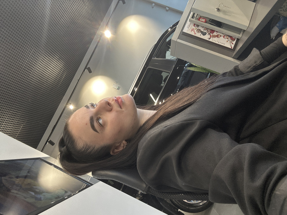 A young adult female with dark hair in a high ponytail, wearing a black blazer, is depicted seated in what appears to be a car dealership showroom. Her profile is visible, with her gaze directed upwards and to the right of the frame. In the background, a black SUV is partially visible, alongside a white wall with a large, dark screen reflecting a blurry image. The ceiling features a gray grid-patterned panel with multiple track lights emitting bright illumination. To the woman's right, an open, light gray safe or cabinet is visible, with a key hanging from its lock. Inside the safe, a vertical holiday-themed advertisement featuring individuals in Santa hats and the "Haval" brand logo is displayed. A green plant is partially visible behind the safe, which rests on a dark counter or desk. The presence of Haval branding indicates the location is a Haval car dealership in Saint Petersburg, Russia.