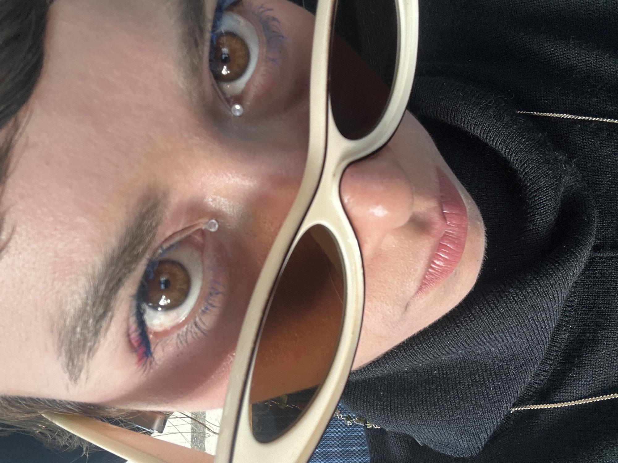 A close-up portrait captures a person's face, oriented approximately 90 degrees counter-clockwise, as they pose in Los Angeles, United States of America. The subject, a young adult, looks slightly upward and to the right, with a pair of cream-colored, retro-style sunglasses held up, partially obscuring the nose and right cheek.
The subject's prominent features include brown eyes, enhanced with blue eyeliner on the lower lash line and a touch of pink or red makeup near the inner corner of the left eye. Dark, well-defined eyebrows frame the eyes. Two small, clear, teardrop-shaped decorative piercings or gems are visible—one below the outer corner of the left eye and another on the skin beneath the right eye. The lower lip, appearing moisturized with a subtle pink tint, is also visible.
The sunglasses feature dark, reflective lenses set in stylish, slightly cat-eye shaped cream frames. A portion of a black, finely ribbed top, possibly a sweater or turtleneck, is seen covering the person's chest and neck area. A thin, delicate silver chain necklace rests against the black fabric. Bright lighting illuminates the scene, creating reflections on the sunglasses and highlights on the skin and eyes, contributing to a chic and artistic aesthetic. No visible text is present in the image.