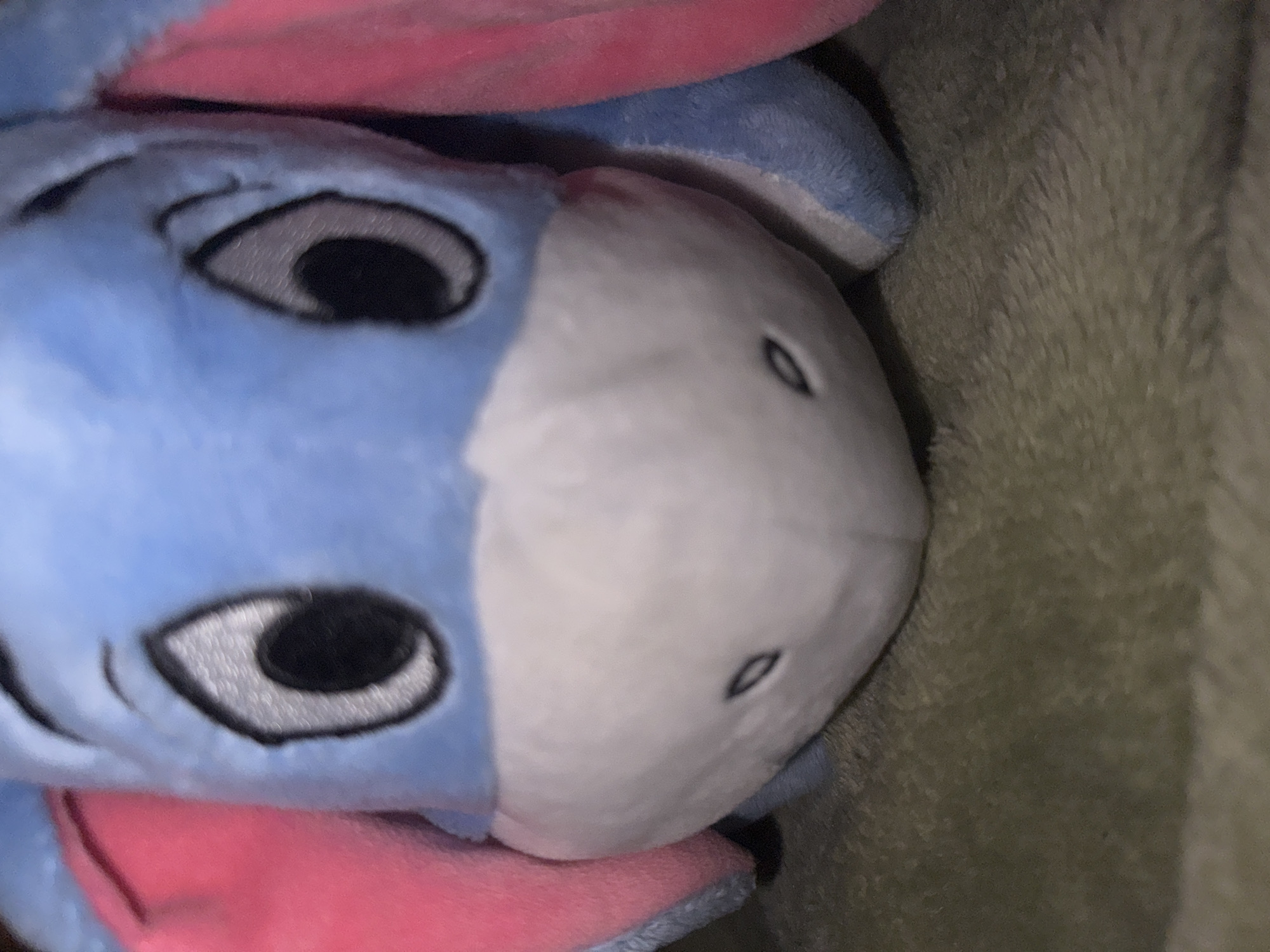 The image is a close-up shot of a stuffed Eeyore toy. The toy is predominantly blue and pink, with large black eyes outlined in gray. Its snout is white with two small, dark nostrils. The toy is lying on a green, fuzzy surface, possibly a blanket or carpet. The lighting is dim, suggesting an indoor setting. There is no visible information in the image to indicate its location in Burgas, Bulgaria.