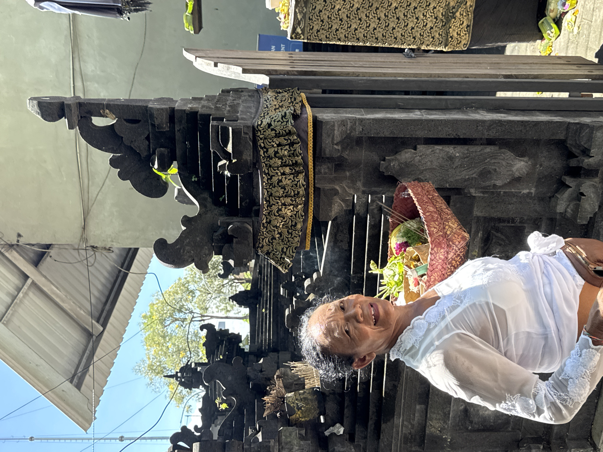 An older woman, likely Balinese, smiles warmly at the camera while standing in front of a dark, intricately carved stone structure, possibly part of a temple or shrine. She has short, dark hair with visible grey streaks. She is dressed in traditional attire, including a white, semi-sheer top with embroidered sleeves and a sarong or wrapped garment around her waist. In her arms, she carries a woven basket filled with colorful offerings, such as flowers, leaves, and fruit, and wisps of smoke, likely from incense, rise near them.
The stone structure behind her is adorned with patterned gold and black fabric, characteristic of Balinese religious sites. Above the main structure, a wooden platform is visible. In the background, under a bright blue sky, are green trees, a corrugated metal roof, and power lines, suggesting an urban or semi-urban outdoor setting during daylight hours. A partial blue sign with the letters "ANT" and "ENT" is faintly visible in the background, possibly from a "RESTAURANT" or similar establishment. The scene conveys a sense of cultural and religious activity in Denpasar, Indonesia.