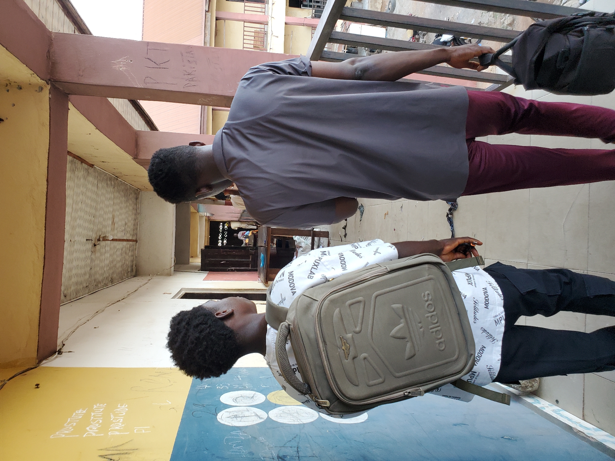 The image captures two young men standing on a balcony in what appears to be a building under renovation in Mararaba, Nigeria. The man closer to the camera wears a white shirt with "MODOVA" printed across it, black pants, and has a grey Adidas backpack. He is standing next to a wall painted with blue and yellow sections, with chalk writing and drawings on the blue portion. The second man wears a grey short-sleeved shirt and maroon pants, holding a black bag and looking down. The building is old and somewhat dilapidated, with unfinished plastering and exposed brickwork. Graffiti is visible on the support beams. The balcony overlooks a street with other buildings in the background.