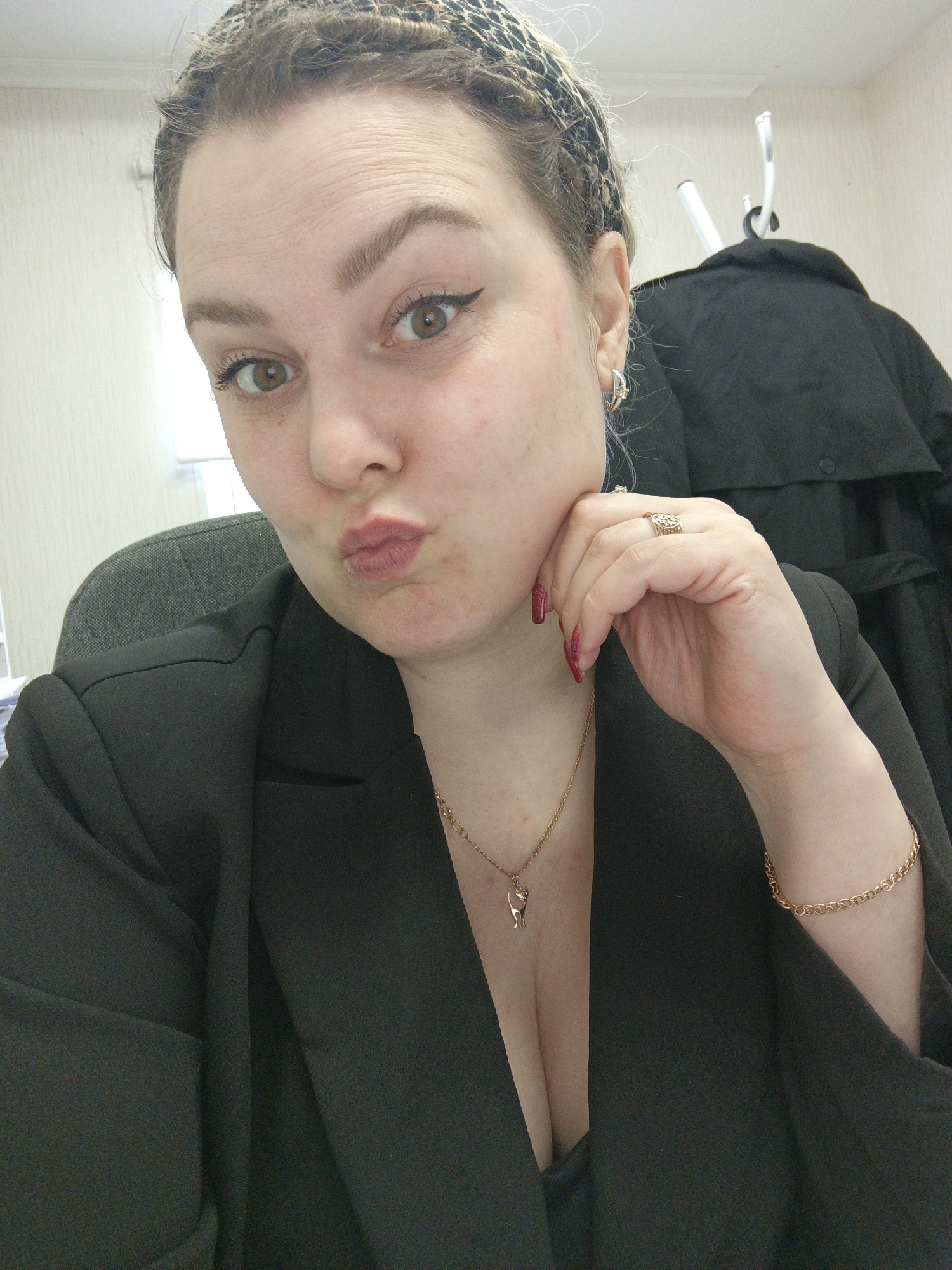 The image displays a close-up portrait of an adult female in the city of Noviy, Russia, captured from the chest up. The subject has light brown hair styled with visible braids or twists, partially covered by a dark, patterned headband. Her eyes are light brown/hazel, and she wears black winged eyeliner and defined eyebrows. Her lips are pursed in a "kiss" gesture, and her right hand, with dark red painted fingernails and a gold-colored ring on the ring finger, is positioned against her chin. A gold-colored chain necklace with a small, stylized cat-shaped pendant is visible around her neck, and a gold-colored chain bracelet is present on her left wrist. She is dressed in a black blazer over a dark, V-neck undergarment. The background consists of light-colored, possibly paneled, walls. A section of a dark grey upholstered office chair is visible to the left of the subject. To the right, a white coat rack holds a black garment. The scene is illuminated by indoor lighting.