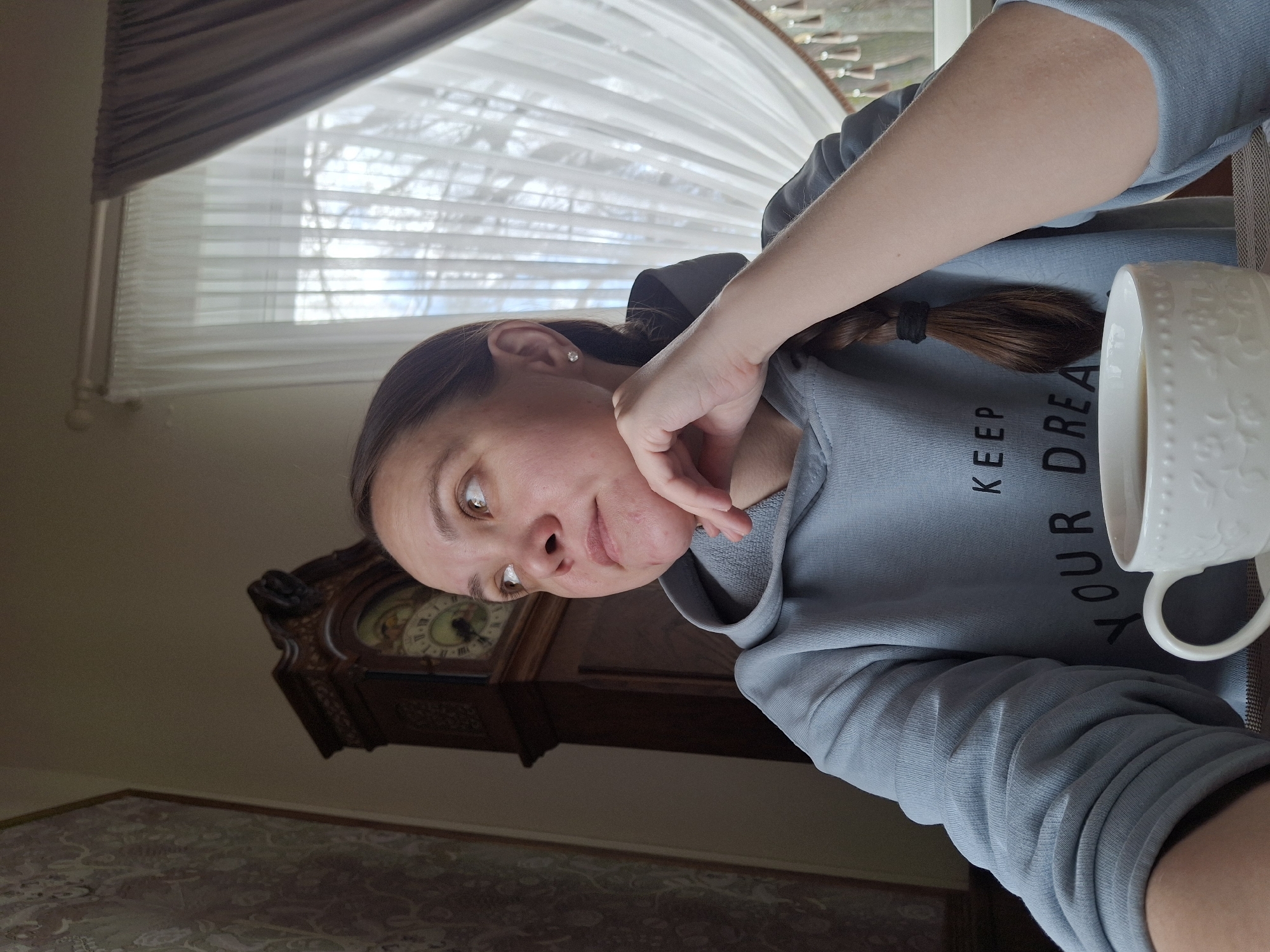This indoor image, taken from a slightly rotated perspective, features a female subject in what appears to be a casual moment. She has brown hair tied back with a black hair tie and is wearing a light blue-grey hooded sweatshirt. A small stud earring is visible in her right ear. Her brown eyes are directed to her left, and her hand rests thoughtfully on her chin, conveying a pensive or calm expression.
A white ceramic mug with an embossed floral or decorative pattern is positioned to her right, containing a light-colored liquid, possibly a warm beverage. Partially visible text on her sweatshirt reads "KEEP YOUR DREA," suggesting "KEEP YOUR DREAMS."
The background reveals a brightly lit room, likely a living area, with natural light entering through a large window. The window is adorned with white horizontal blinds and a dark grey fabric valance. Outside, the bare branches of trees against a bright sky suggest a colder season, such as late autumn or winter. To the subject's left, a prominent dark wooden grandfather clock with ornate carvings and a classic Roman numeral clock face adds a traditional touch to the decor. A patterned rug or carpet is also partially visible in the lower foreground. The scene depicts a quiet, reflective daytime moment within a home in Sarapul, Russia.