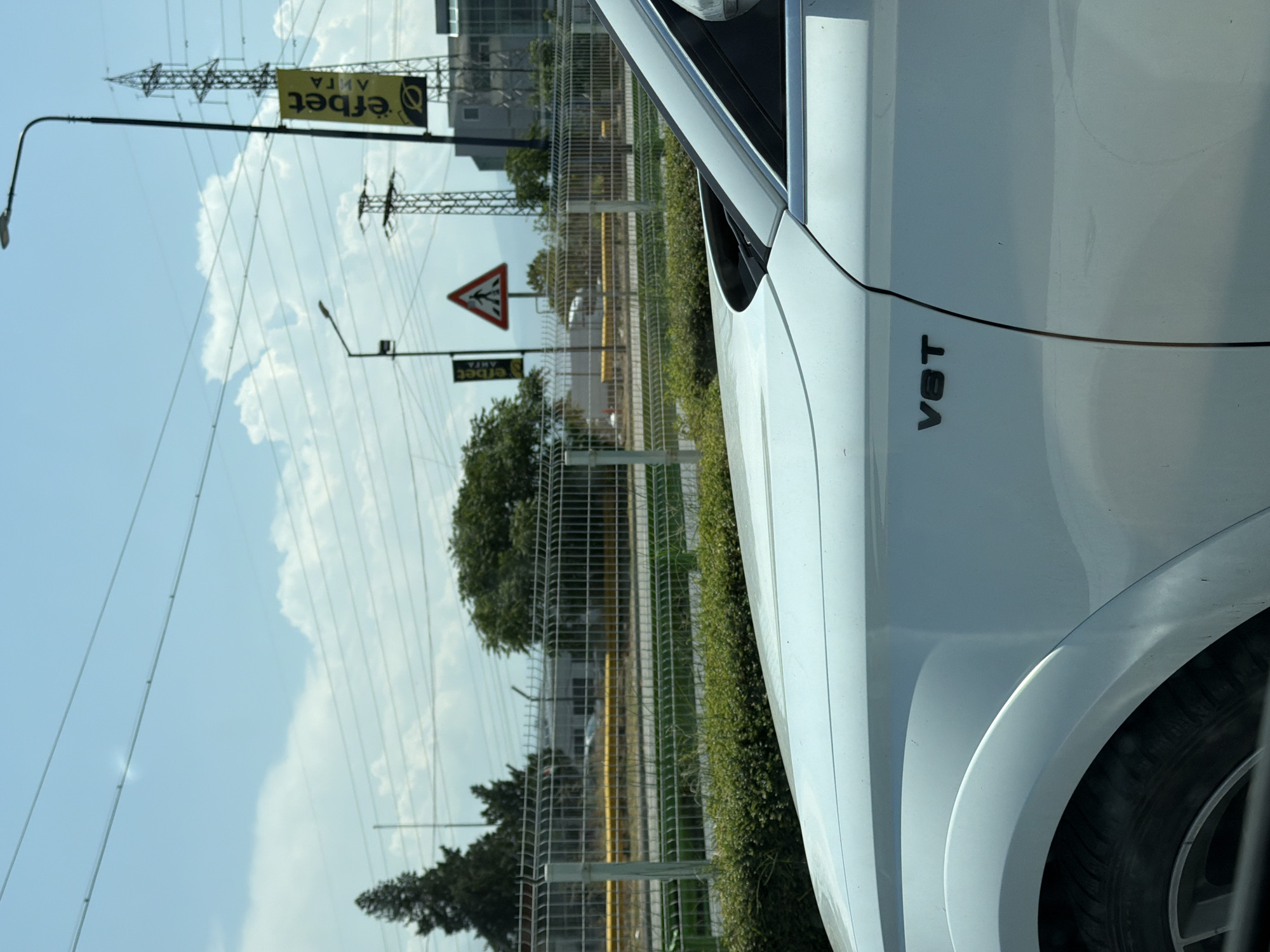 The image shows the side of a white car, with a "V8T" emblem visible on the door. In the background, there is a chain-link fence, some greenery, and trees. Beyond the fence, there are buildings, utility poles, and power lines. A yellow banner with "efbet" printed on it hangs from a pole. A pedestrian crossing sign is also visible. The scene suggests an urban or suburban environment, likely near a road or industrial area. The image was taken on a clear day, with a blue sky and some clouds. The location is Pazardzhik, Bulgaria.