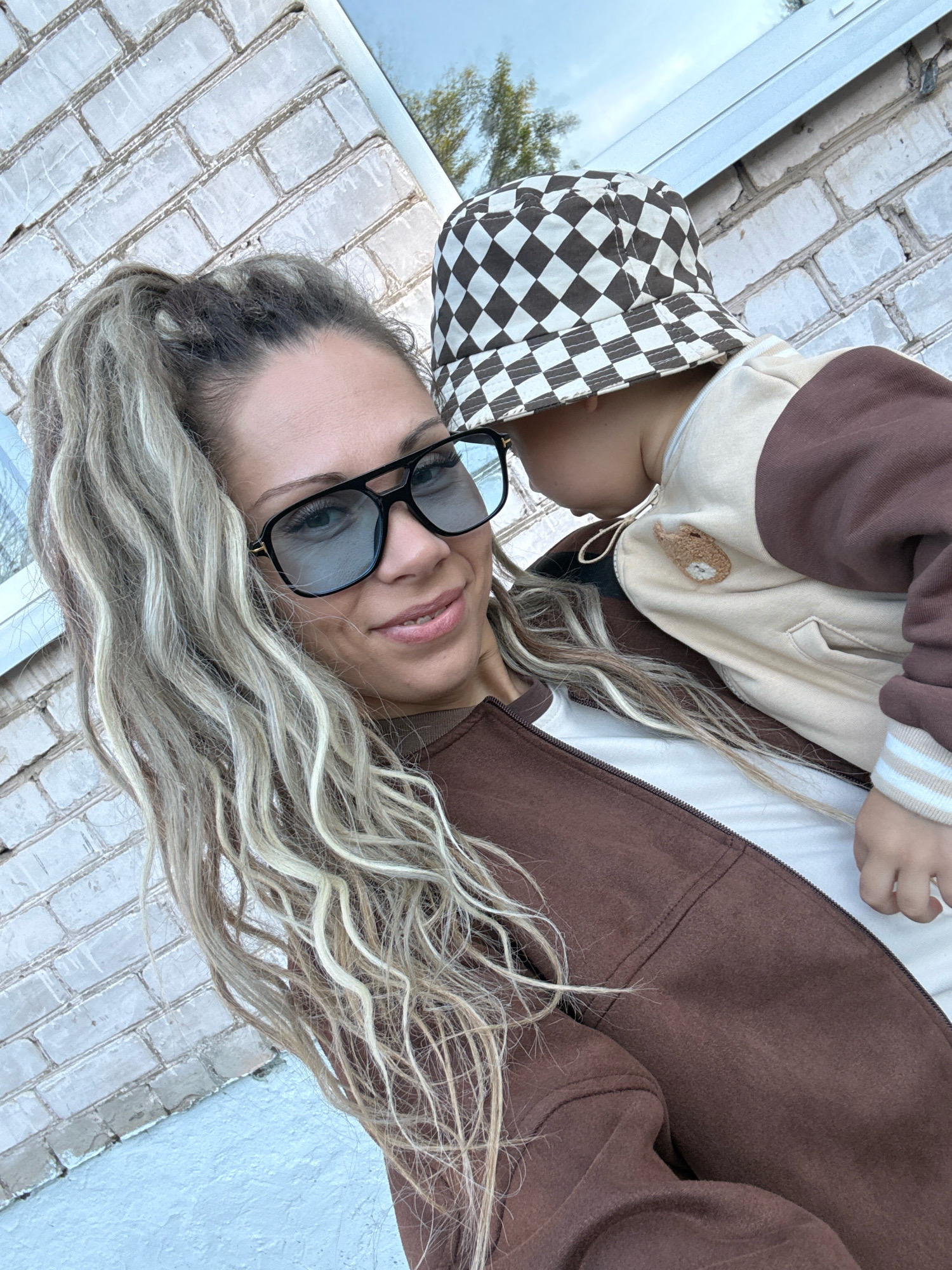 A woman and a child pose for a selfie in front of a white brick wall, likely in Rusanovo, Russia. The woman, with blonde hair pulled back in a ponytail and large black-rimmed glasses, smiles at the camera. She wears a brown jacket over a white shirt. The child, wearing a brown and white checkered bucket hat and a cream-colored jacket with brown sleeves, is held by the woman, facing away from the camera. The background consists of the brick wall and a hint of sky and tree branches.