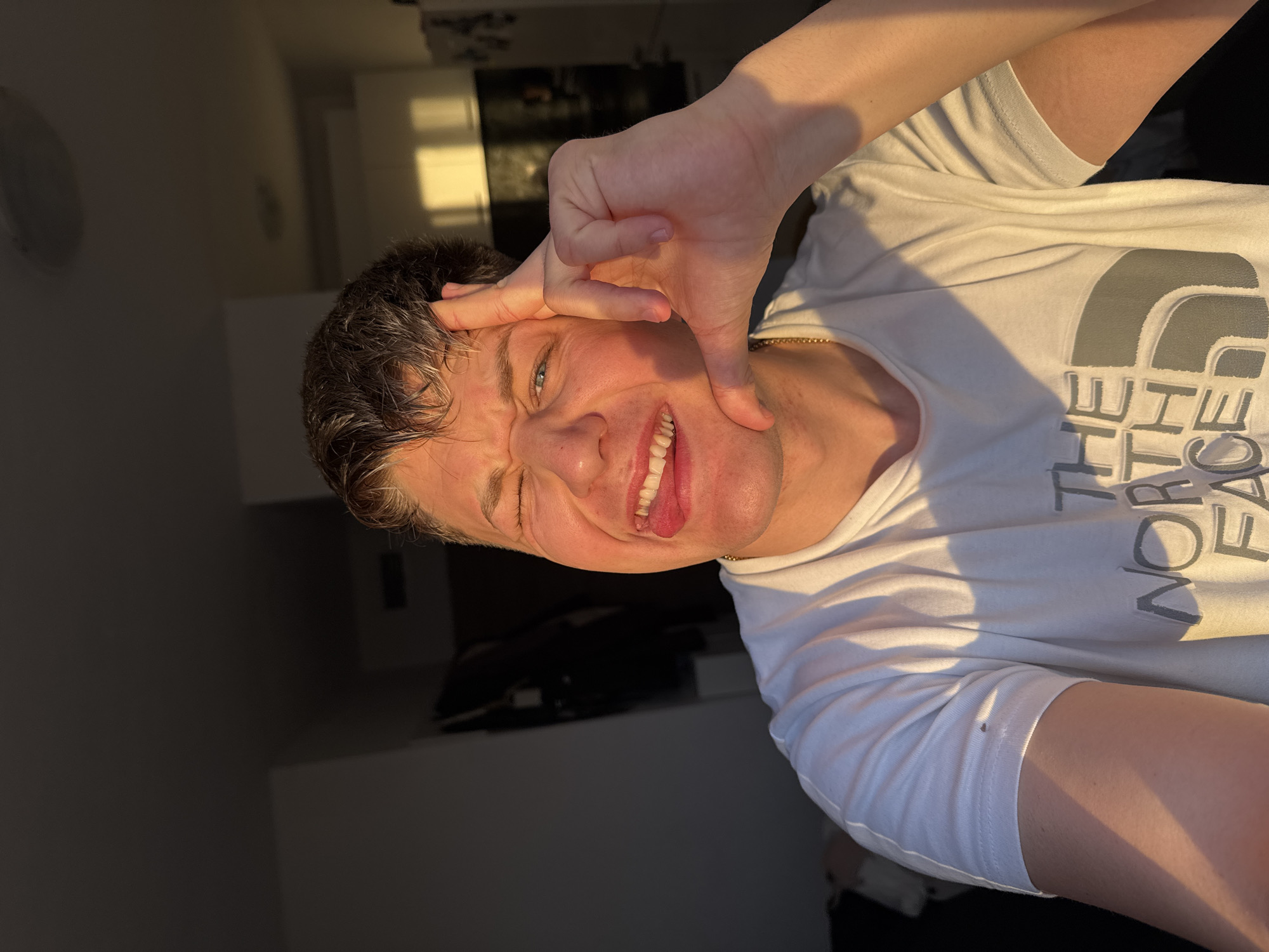 A person is posing for a photo indoors, wearing a white t-shirt with "THE NORTH FACE" printed on it. The person is making a face and has their hand on their face, forming a square-like shape around their eye and cheek. The background is blurry and indistinct, but appears to be a room with some furniture and possibly a window with sunlight coming through. The person is wearing a gold chain around their neck. The image does not provide information to determine the location in Plovdiv, Bulgaria.