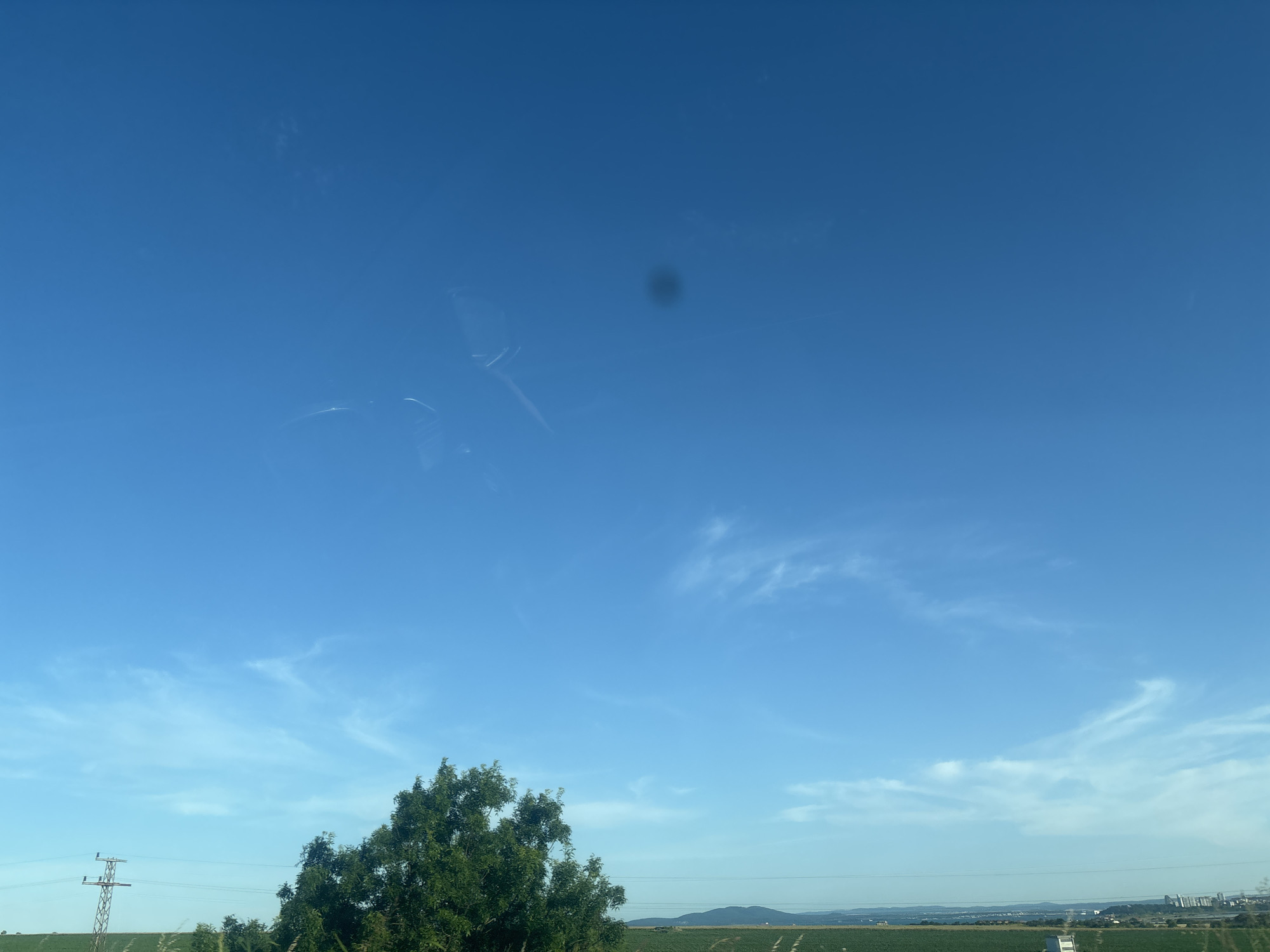 A wide shot captures a vast expanse of clear blue sky, with wispy white clouds scattered across its surface. A small, dark circular blemish is visible in the upper center of the sky. The lower portion of the image shows a distant green landscape, likely agricultural fields, stretching towards a horizon dotted with hills and some low-rise buildings in the far distance. To the left, a solitary green tree with dense foliage stands prominently, and further left, a tall, grey electrical pylon with power lines extends across the frame. The overall impression is of a rural or semi-rural setting under a bright, clear sky. This image could be of the Burgas region of Bulgaria, as the landscape and infrastructure are consistent with the area.