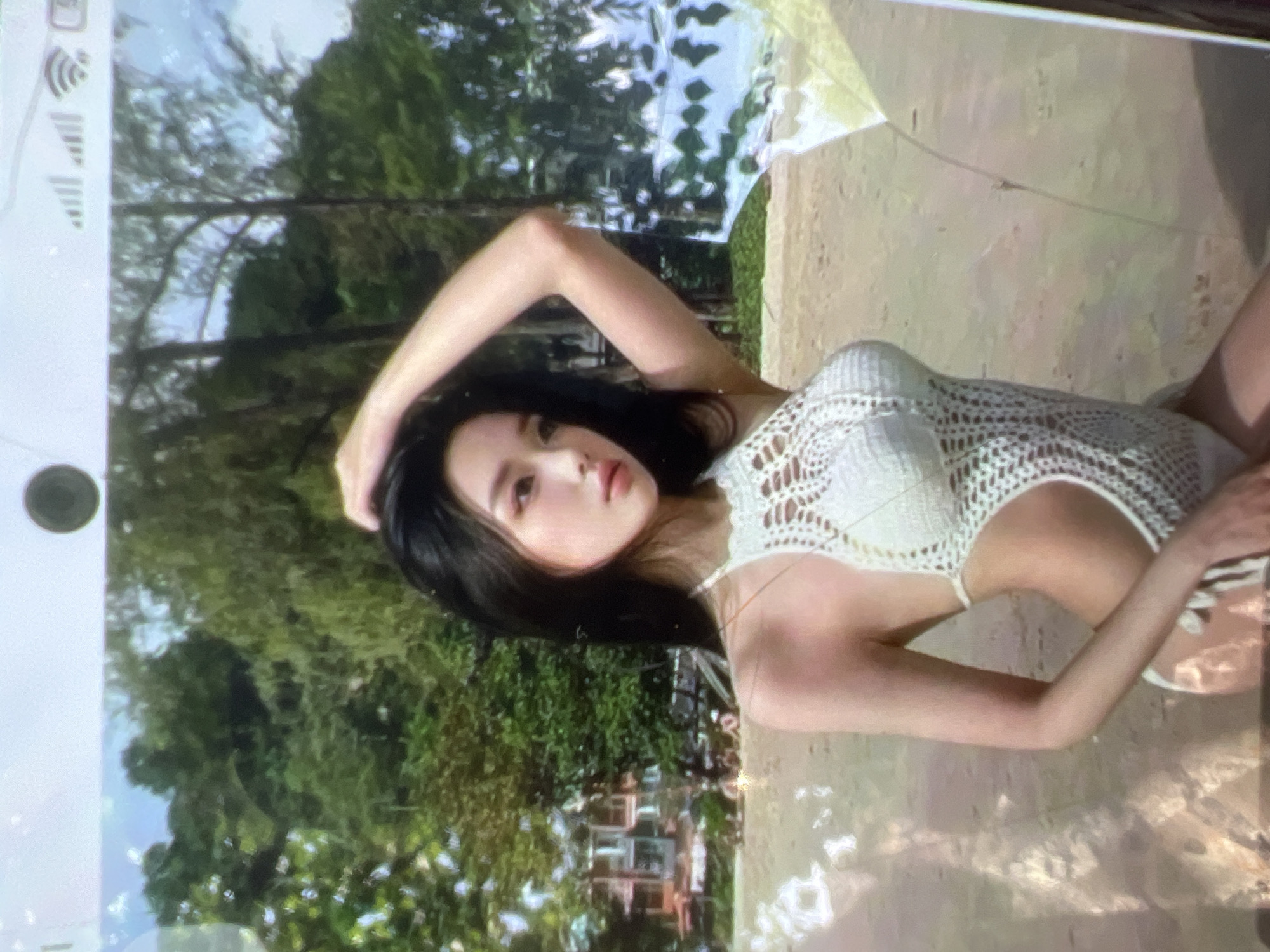 A young woman with long, dark hair is posing on a sandy beach. She is wearing a white, crocheted one-piece swimsuit with a halter neckline and cut-outs on the sides. Her left arm is raised and bent behind her head, and her right arm is down by her side. She is looking towards the viewer with a neutral expression. The background shows lush green trees, a few buildings that appear to be hotels or resorts, and the ocean. The sand is light brown and appears wet in some areas, suggesting the tide is close. The lighting indicates it is daytime, likely sunny, with some reflections on the water and sand. The scene is likely a tropical or subtropical location.
