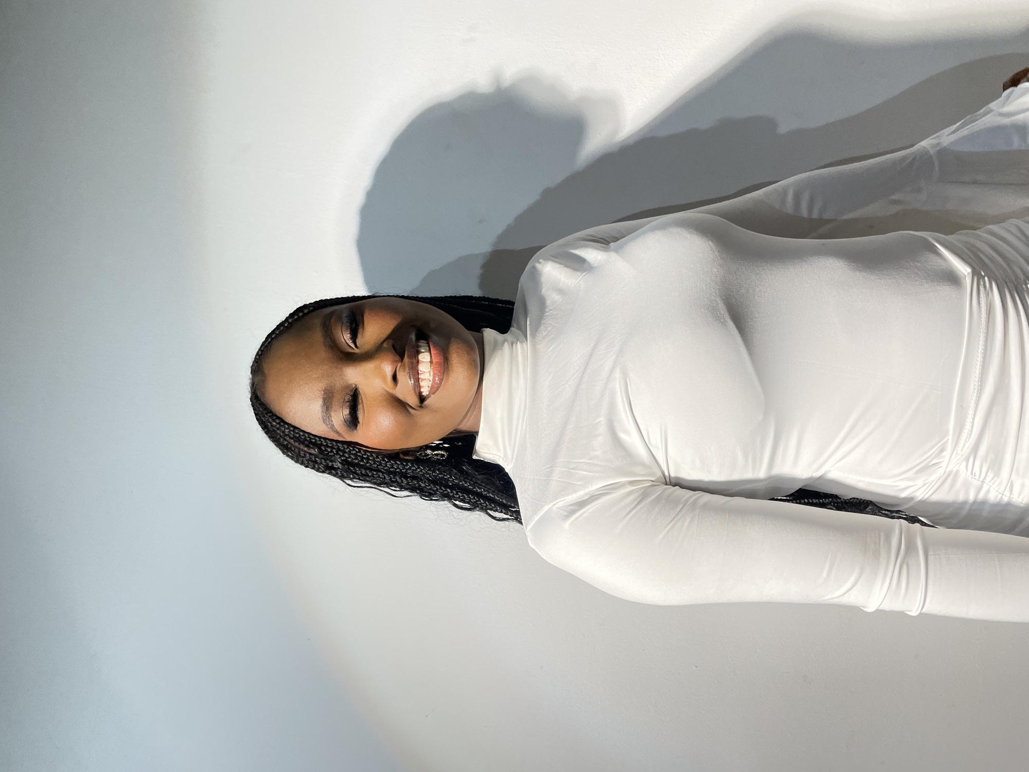 A Black woman is captured in a medium shot, smiling broadly with a joyful expression, her eyes slightly closed. Her dark hair is styled in numerous neat braids. She is wearing a form-fitting, high-necked, long-sleeved white garment, which could be a dress or a top. The fabric of her attire shows subtle drapes and folds.
The setting appears to be an indoor space with a plain white wall as the background. Strong, directional lighting, possibly from an artificial source or a window, casts prominent, soft-edged dark grey shadows on the wall behind and to the side of the woman. These shadows suggest a light source positioned to her left (viewer's right). The overall lighting is bright, emphasizing the white of her clothing and the details of her face.
Her pose is upright, and her smile conveys happiness and warmth. There are no other discernible objects or details in the minimalist background. No text is visible within the image. While the image itself does not contain specific location cues, it is contextualized as being from Barakin Akawo, Nigeria.