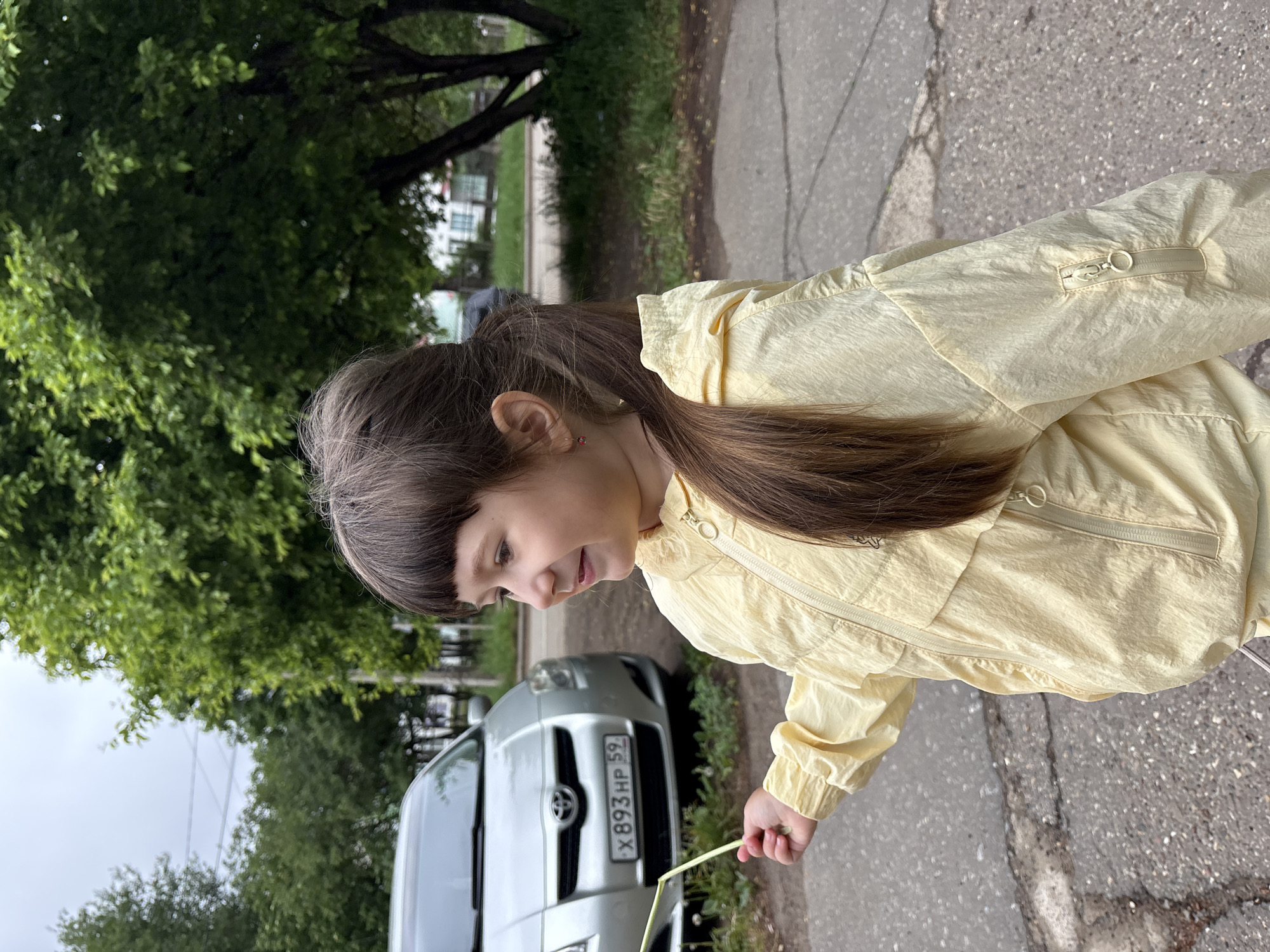The image shows a girl with brown hair and bangs wearing a yellow jacket, standing on a grey paved surface. She appears to be holding a yellow straw or stick. The background features a silver car, trees, and a glimpse of a building. The car's license plate reads "X 893 HP 59," indicating the Perm region of Russia. While it's difficult to precisely pinpoint the location, based on the car registration and the context provided in the prompt, the setting can be considered Chaikovsky, Russia.