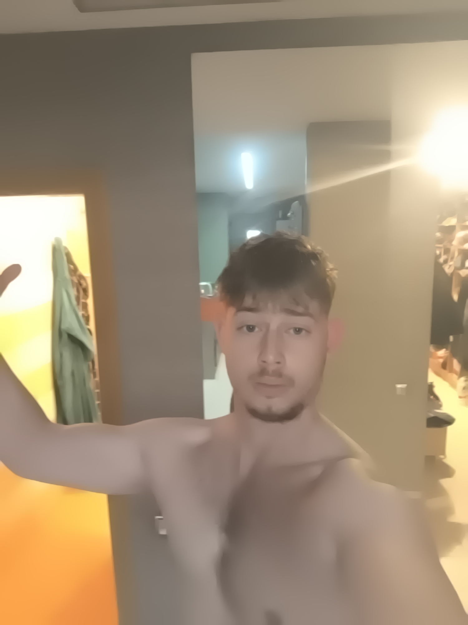A shirtless, fair-skinned man with short brown hair and a beard stands indoors. The background is blurry, revealing a hallway with a partially visible wardrobe and interior design features. The exact location in Sofia, Bulgaria is not determinable from the image.