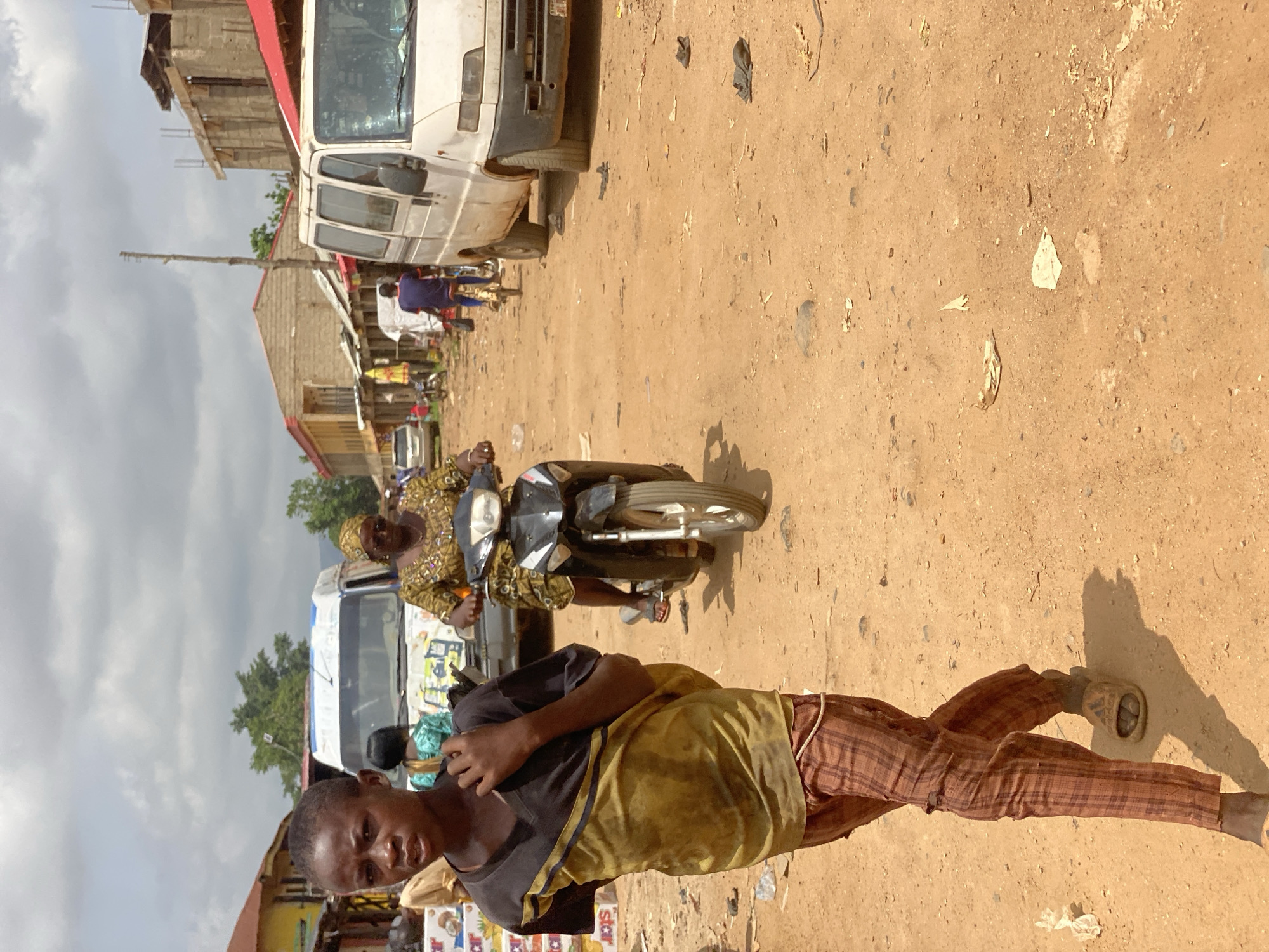 The image shows a street scene in Kafanchan B, Nigeria. A young person wearing a black t-shirt with a yellow fabric, brown plaid pants, and sandals is standing in the foreground. An older woman is riding a motorcycle directly behind this person. A white van is parked on the side of the road. The ground is brown and dusty, and there are buildings and people in the background. The sky is overcast.