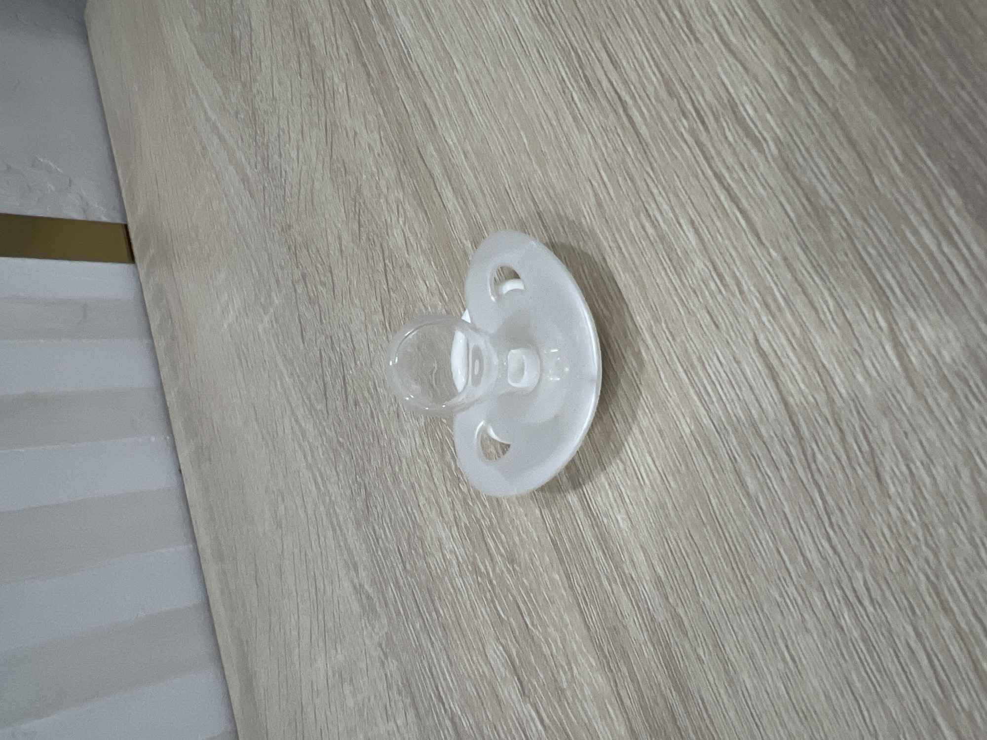 A white pacifier with a clear nipple is resting on a light-colored, wood-grained surface. The background consists of a wall with alternating vertical stripes of light and dark grey, and a sliver of a gold-colored vertical element. The lighting is soft, casting faint shadows. The scene is static, with no people or discernible actions. The setting appears to be indoors, likely a domestic environment. The location is not specified as Veteran-2, Russia.