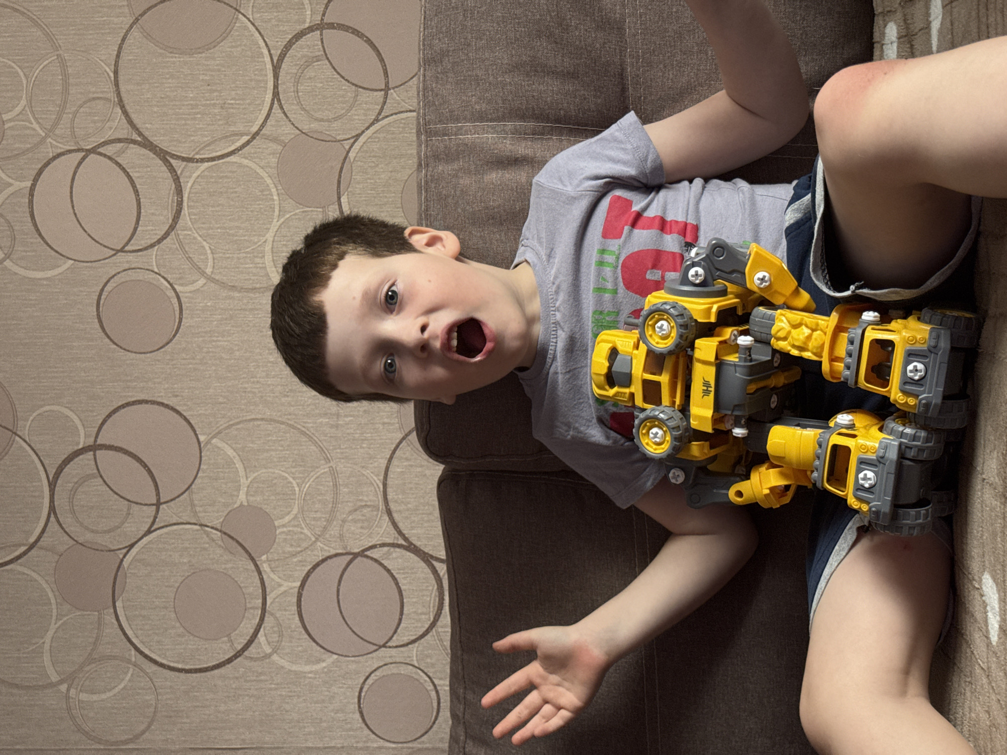 A young boy lies on a brown couch in front of a wall with a circular patterned wallpaper. He is wearing a gray t-shirt and blue shorts. A yellow and gray toy construction vehicle is positioned on top of him. His mouth is open, and he appears surprised or expressive. The image was taken indoors, likely in a private residence, but the location Кемуль, Russia is irrelevant.