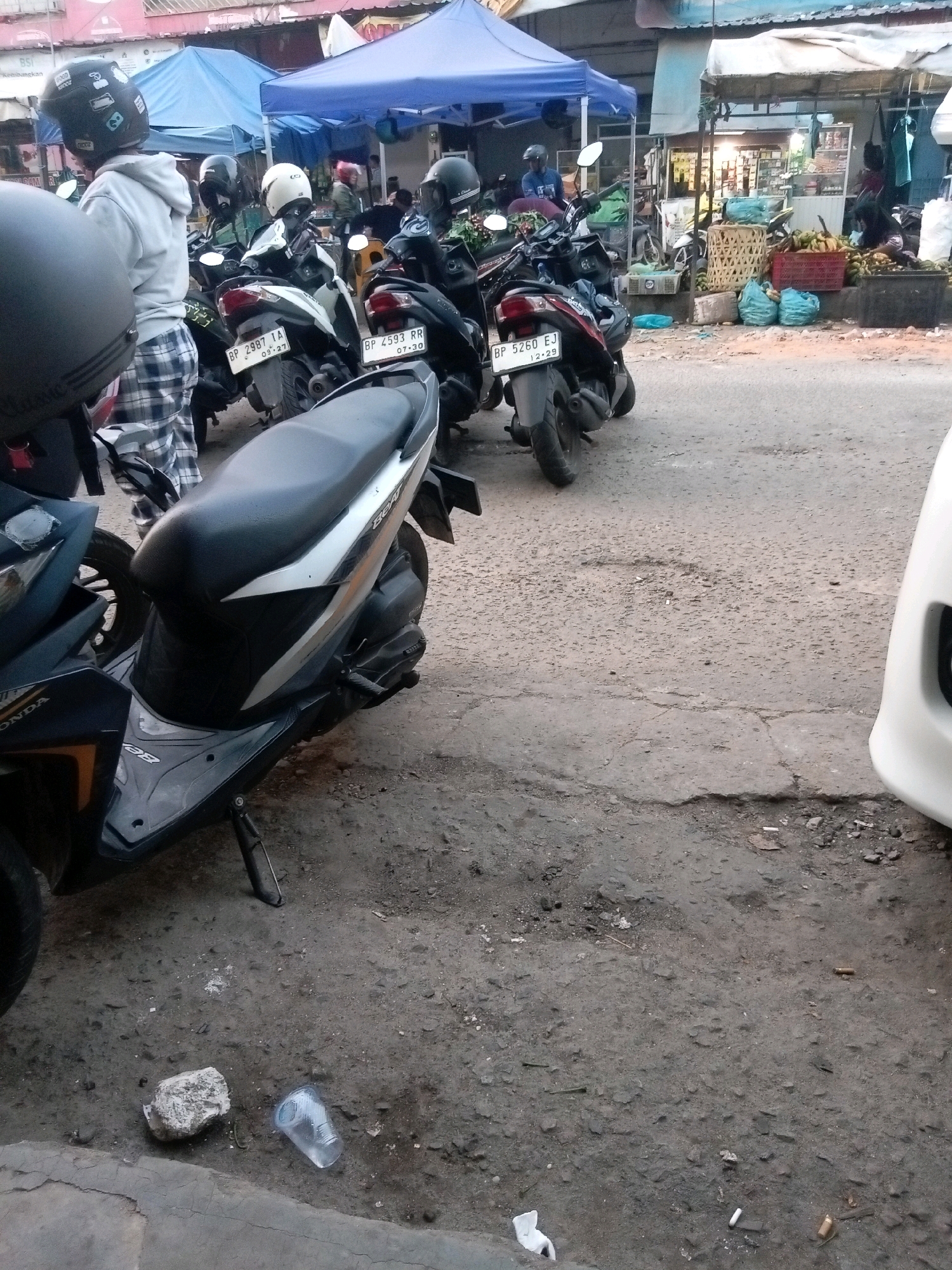 This is an eye-level, medium shot capturing a street scene in Batam, Indonesia, likely near a market or outdoor vendor area, with an emphasis on parked motorcycles and the ground. In the foreground, a black and silver scooter, identified by "HONDA" and "BEAT" on its body, is parked on the right side of the frame. To the left, a black motorcycle helmet with various stickers is partially visible. A person wearing a white hoodie and plaid pants stands behind this helmet, facing away from the camera.
Behind the foreground scooter, several other motorcycles are parked, their license plates indicating them as follows: "BP 2987 IA", "BP 2593 RR", and "BP 5260 EJ". These motorcycles are positioned in front of stalls covered by blue and white tarps, suggesting a commercial or market environment. In the background, stalls are visible with various goods, including crates of produce like bananas, and blue plastic bags. A few people are faintly discernible in the background, some appearing to be vendors or customers.
The ground in the scene is rough asphalt and dirt, littered with debris including cigarette butts, a white plastic cup, and a piece of concrete. The overall lighting suggests it is daytime, possibly late afternoon or early morning, with soft, diffused light. The scene conveys a sense of everyday life in a busy urban area of Batam.