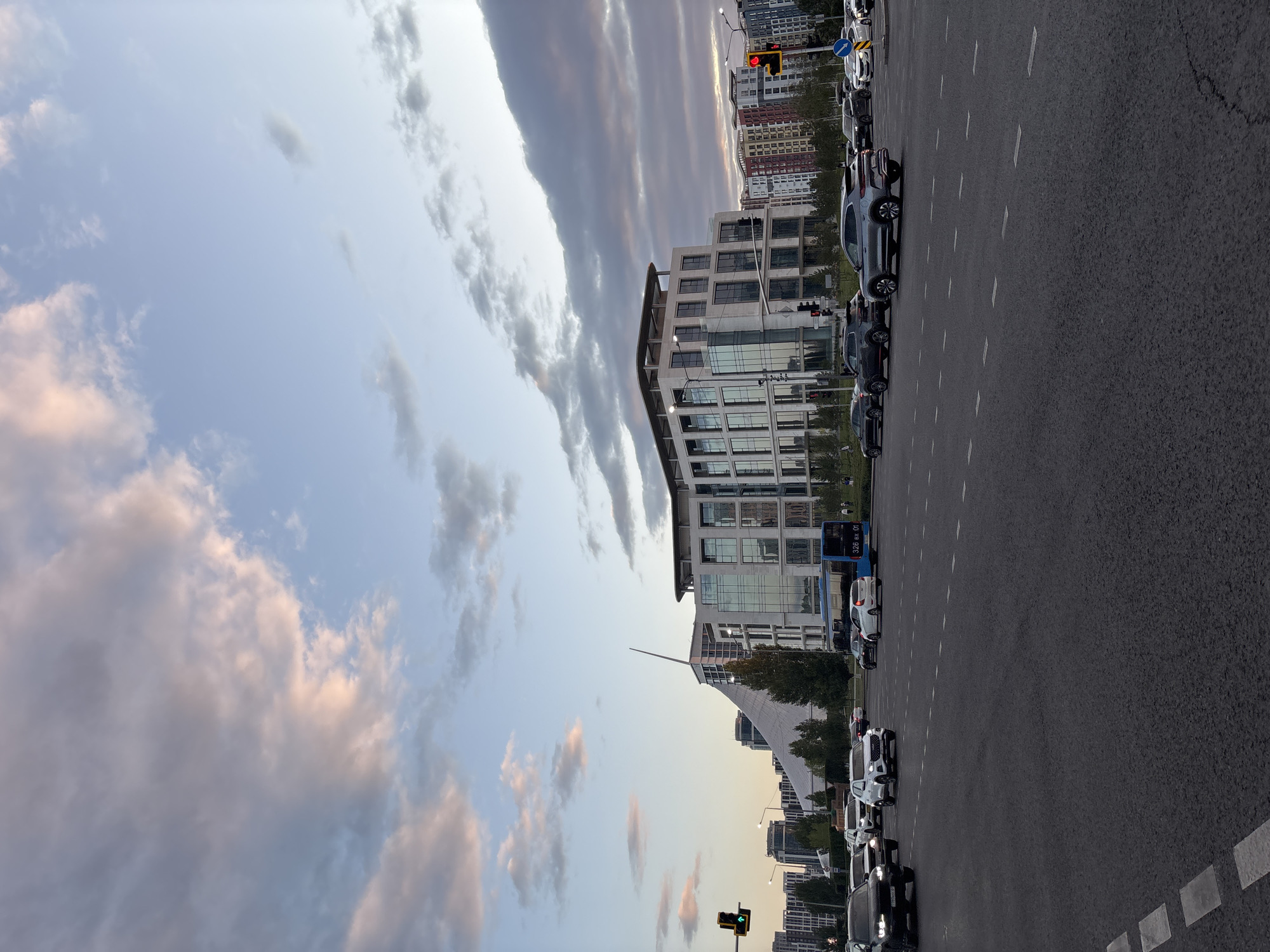 The image captures a wide-angle view of a cityscape in Astana, Kazakhstan, likely during the late afternoon or early evening given the lighting and cloud formations. The foreground is dominated by a multi-lane asphalt road with visible white lane markings and a pedestrian crossing. Several vehicles, including SUVs and sedans, are traversing the intersection, with some stopped at a traffic light. One traffic signal shows a green upward arrow, indicating a permitted movement.
In the mid-ground, a modern, multi-story building with a glass facade stands prominently. To its left, a distinctive white, tent-like structure with a sharp apex rises against the sky. This structure is likely part of a significant architectural landmark in Astana. Adjacent to these buildings are rows of residential buildings with colorful facades. A blue bus is visible on the road, with some passengers potentially inside. Green trees line the area between the road and the buildings, adding a touch of nature to the urban landscape.
The sky above is a mix of pale blue and scattered clouds, some of which are tinged with pink and orange, suggesting the setting sun. The overall impression is one of a busy urban environment during a transitional time of day, showcasing modern architecture and city life in Astana.