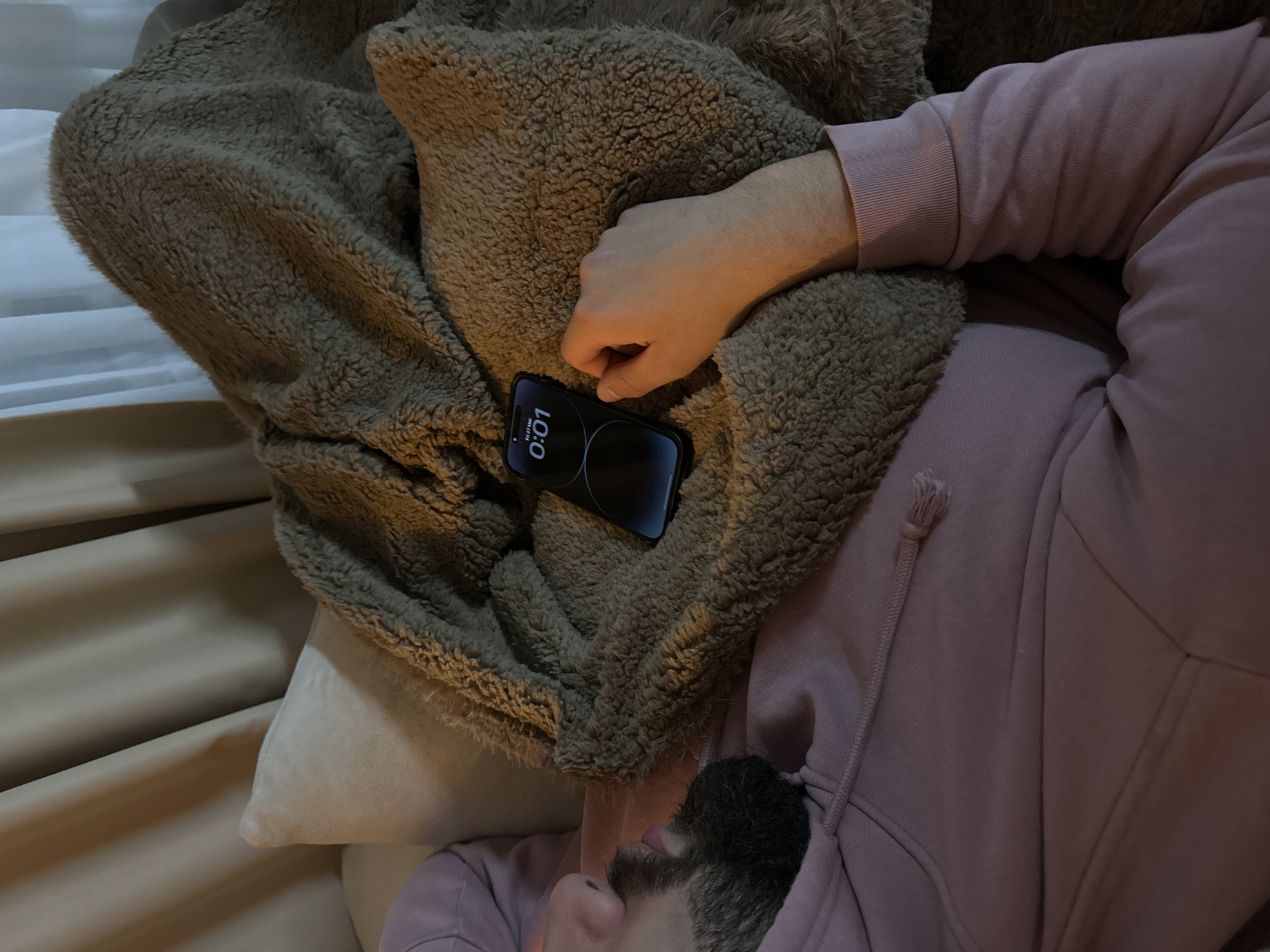 An overhead close-up image depicts a male subject resting indoors. The specific location within Sofia, Bulgaria, cannot be determined from the visual information provided.
The subject, seen from the chest up and facing left, is lying on a light-colored surface, possibly a sofa or bed. He possesses dark facial hair, including a beard and mustache. He wears a light pink or mauve hooded sweatshirt with a visible drawstring and ribbed cuffs on the right sleeve. A brown, textured, sherpa-style blanket or throw covers his torso and lap.
To the subject's right, a dark-colored smartphone, identified as an iPhone, rests on the brown blanket. Its screen is illuminated, displaying "Fri 27 Mar" and the time "0:01". The subject's right hand is visible just below the phone, resting palms down on the blanket, with fingers lightly curled. The hand's skin tone appears fair.
In the background, a light-colored, possibly cream or beige, pillow is visible behind the subject's head. Further behind, vertical drapes or curtains of a similar light hue are partially visible, suggesting an indoor setting with natural light control. The overall lighting is dim.