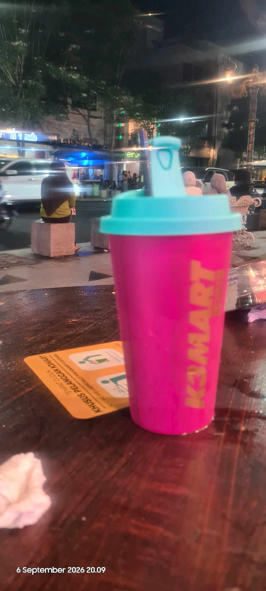 The image depicts an outdoor scene at night in what appears to be a city environment, potentially Surabaya, Indonesia, given the presence of Indonesian text on a placard. In the foreground, a bright pink tumbler with a light blue lid is prominently displayed, tilted slightly and resting on a wooden surface, possibly a table. The tumbler has gold lettering on its side that reads "KEMART".
To the left of the tumbler, a yellow card or placemat is visible. It also features Indonesian text and graphical icons. The text on the card appears to be instructional, possibly indicating designated seating areas or rules for customers, with phrases like "TEMPAT DUDUK KHUSUS PELANGGAN KEMART" (KEMART Customer Seating Area) and "KHUSUS UNTUK PELANGGAN KEMART" (Especially for KEMART Customers).
In the background, a street scene unfolds. Several individuals are visible. One person, seen from behind and wearing a yellow top and dark headscarf, is seated on a concrete block on a sidewalk. Further in the background, more people are gathered, and vehicles are present, suggesting a busy urban setting. The lighting indicates it is nighttime, with streetlights and building lights illuminating the area. Trees and some architectural structures are also visible, contributing to the urban ambiance. The overall impression is that of a casual dining or gathering spot in a city at night.