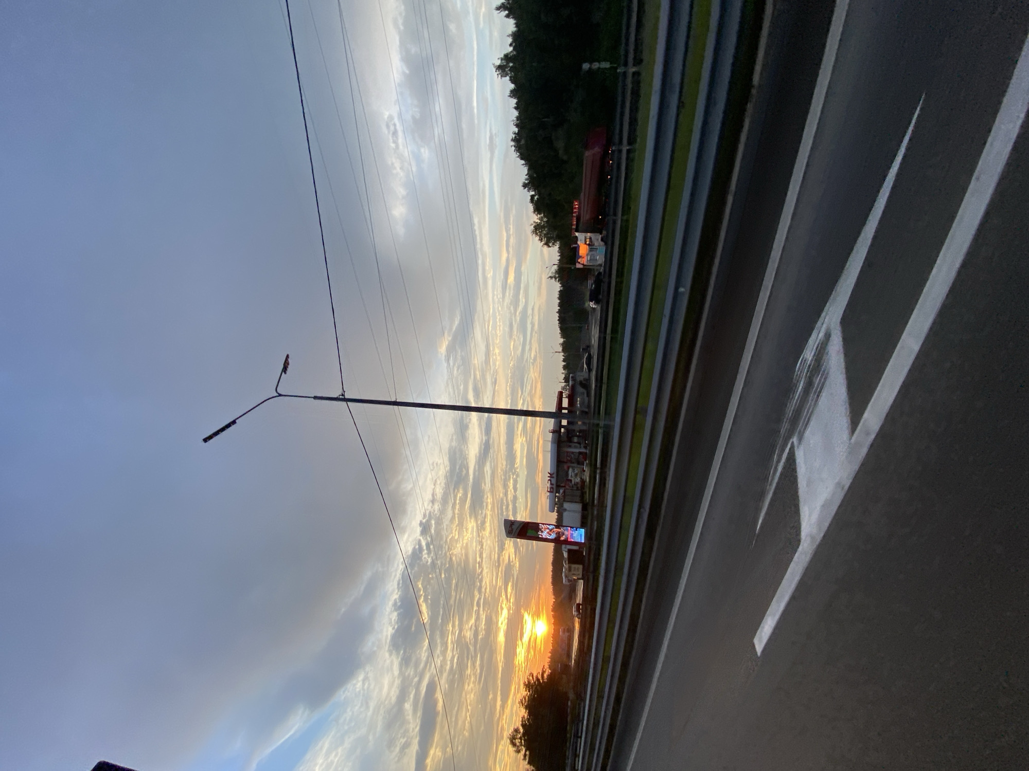 A sunset illuminates a highway in Mamony, Russia. The sky is a mix of blue and grey clouds, with the orange sun setting on the left side of the frame. A roadside billboard with colorful advertisements stands near a gas station. Cars and trucks are visible on the highway, some of which are at the gas station. A tall streetlamp stands on the right side of the road. The highway is paved with asphalt and has white lane markings. A metal guardrail separates the highway from the surrounding greenery.